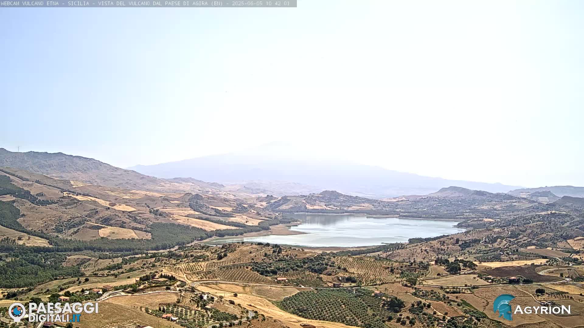 A hazy, sunny day reveals a vast, dry landscape of rolling hills, farmland, and a large lake, with a distant mountain range under a pale sky.