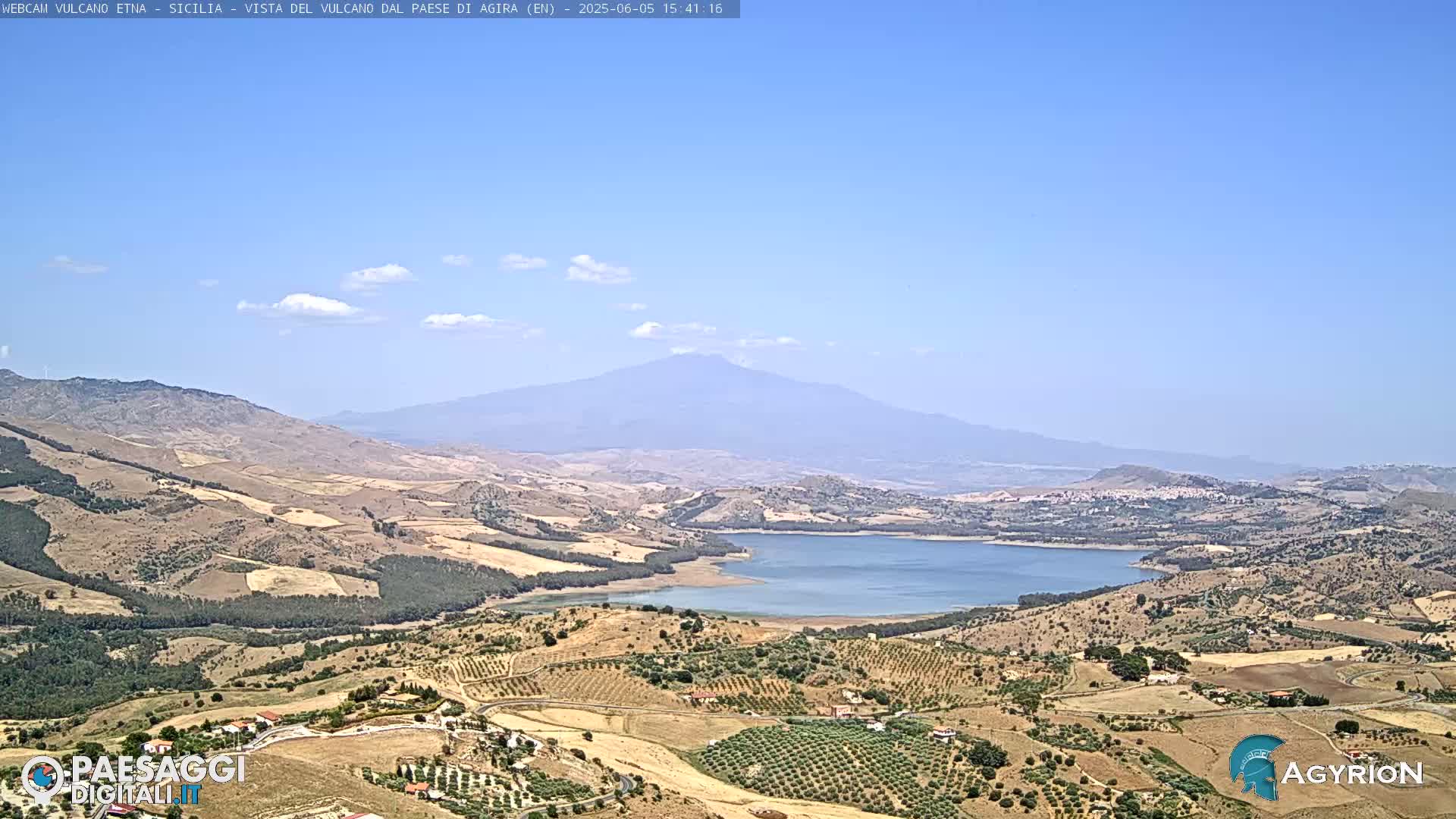 A long shot of a hilly landscape under a clear blue sky shows a large lake and a distant, hazy mountain in the background.