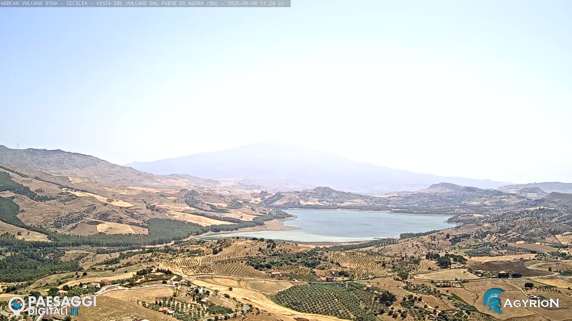 A hazy view shows a large body of water nestled in a valley between rolling hills, with a distant, partially obscured mountain range under a clear sky.