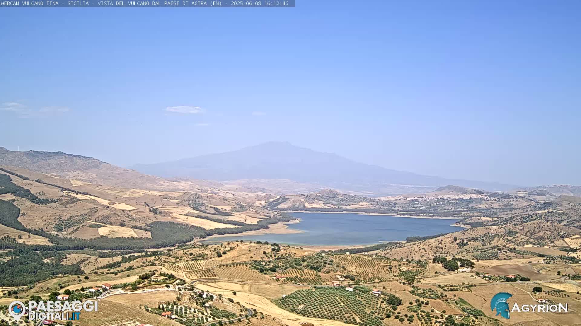 A distant, hazy mountain looms over a dry, hilly landscape dotted with olive groves and a large lake under a clear blue sky.