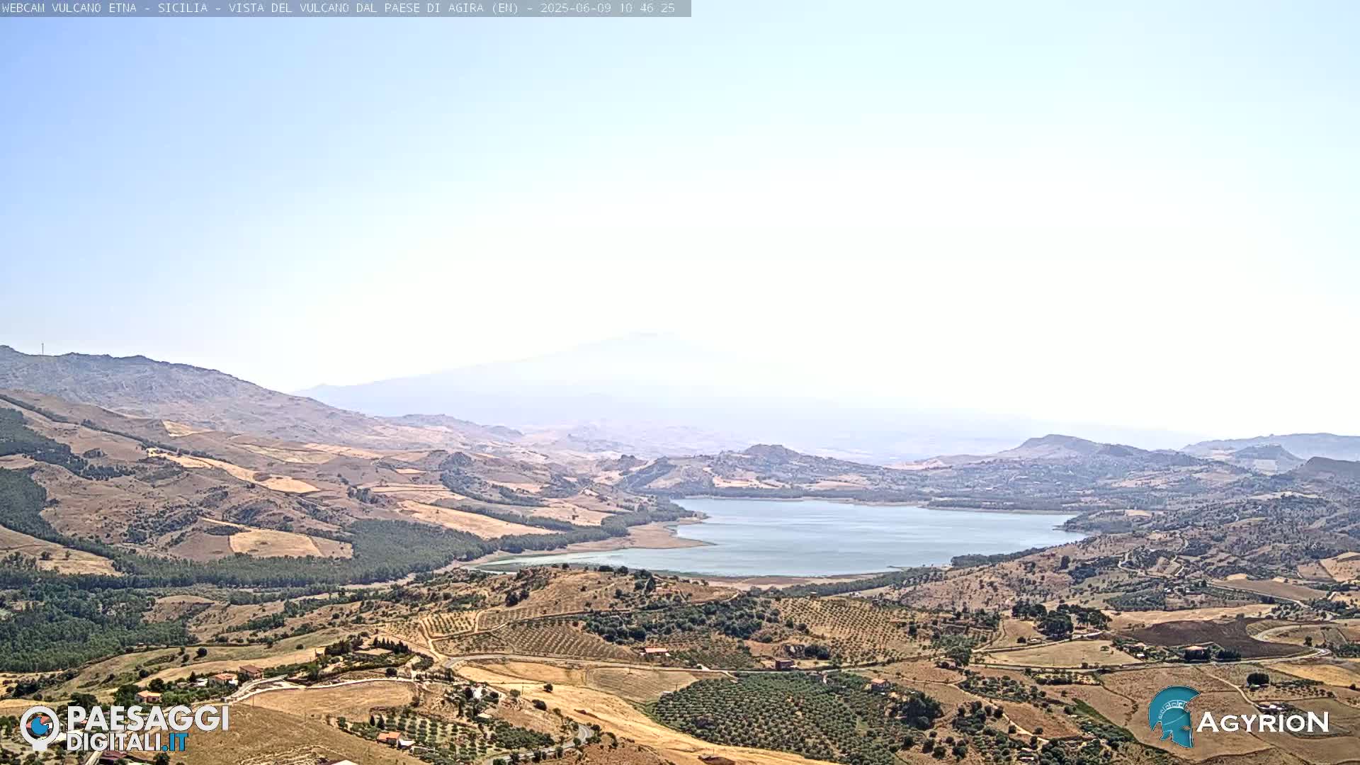 A hazy, sunny day reveals a vast landscape of rolling hills, farmland, and a large lake, with mountains in the distance.