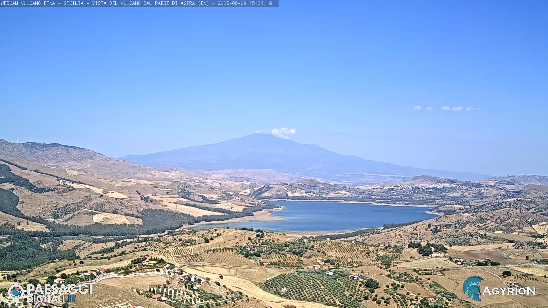 A distant, hazy mountain with a small plume of smoke is visible above a dry, hilly landscape with a lake in the middle distance under a clear blue sky.