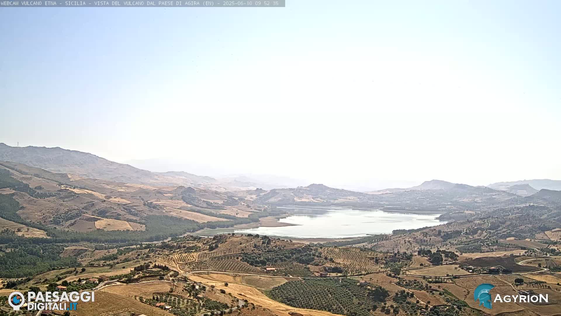 A high-angle, long shot shows a vast, hilly landscape under a hazy, sunny sky, featuring a large lake nestled amongst rolling hills and cultivated fields.