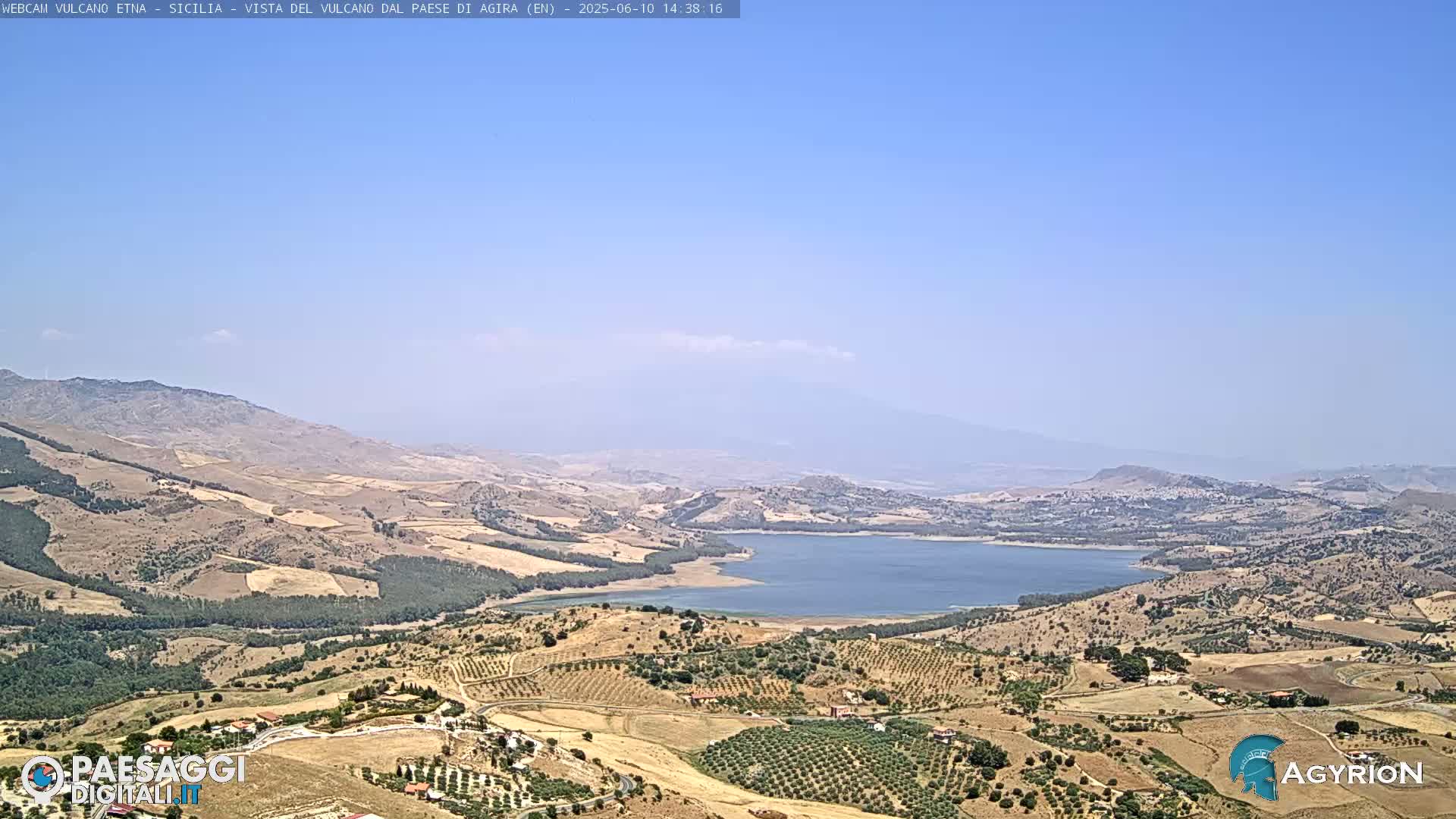 A wide, high-angle view shows a vast, dry landscape under a clear blue sky, featuring a large lake nestled among rolling hills and sparsely vegetated fields.