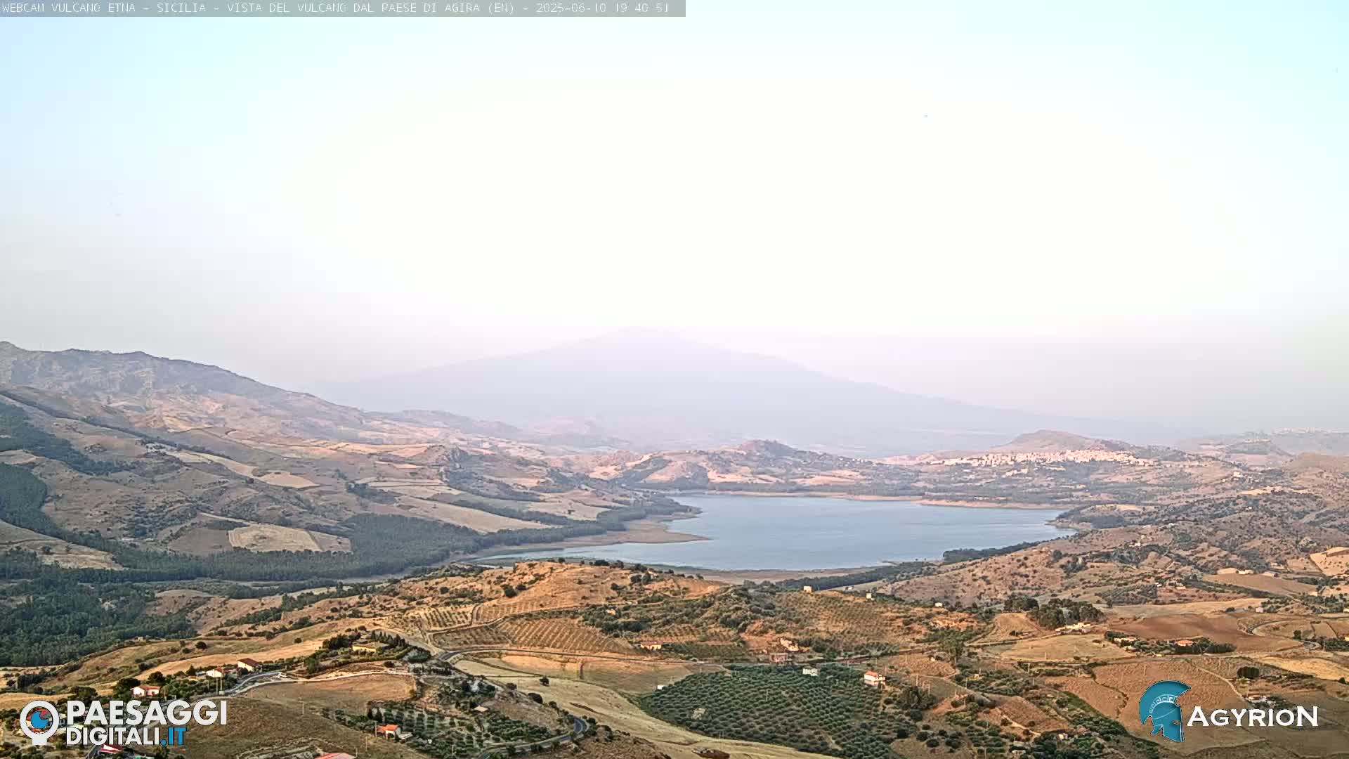 A hazy, long shot of a dry, hilly landscape with a large lake, and a mountain range in the distance under a mostly clear sky.