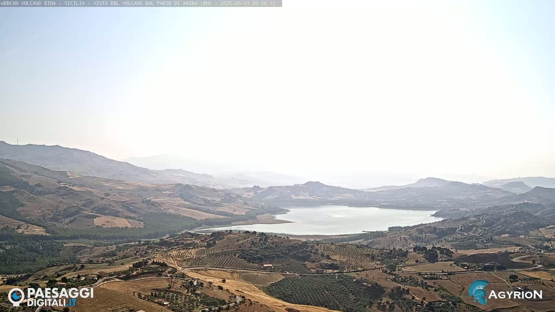 A hazy, sunny view shows a large lake nestled in a valley surrounded by rolling hills and farmland.