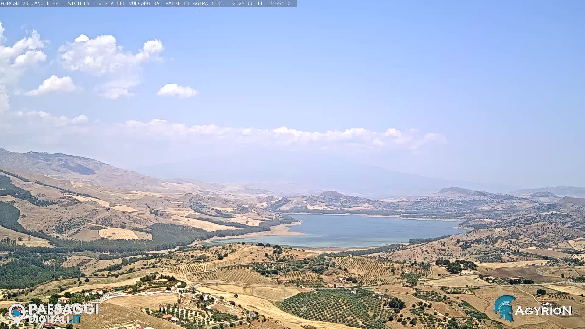 A sprawling landscape under a mostly clear sky with scattered clouds shows a large lake nestled among dry, rolling hills and fields.