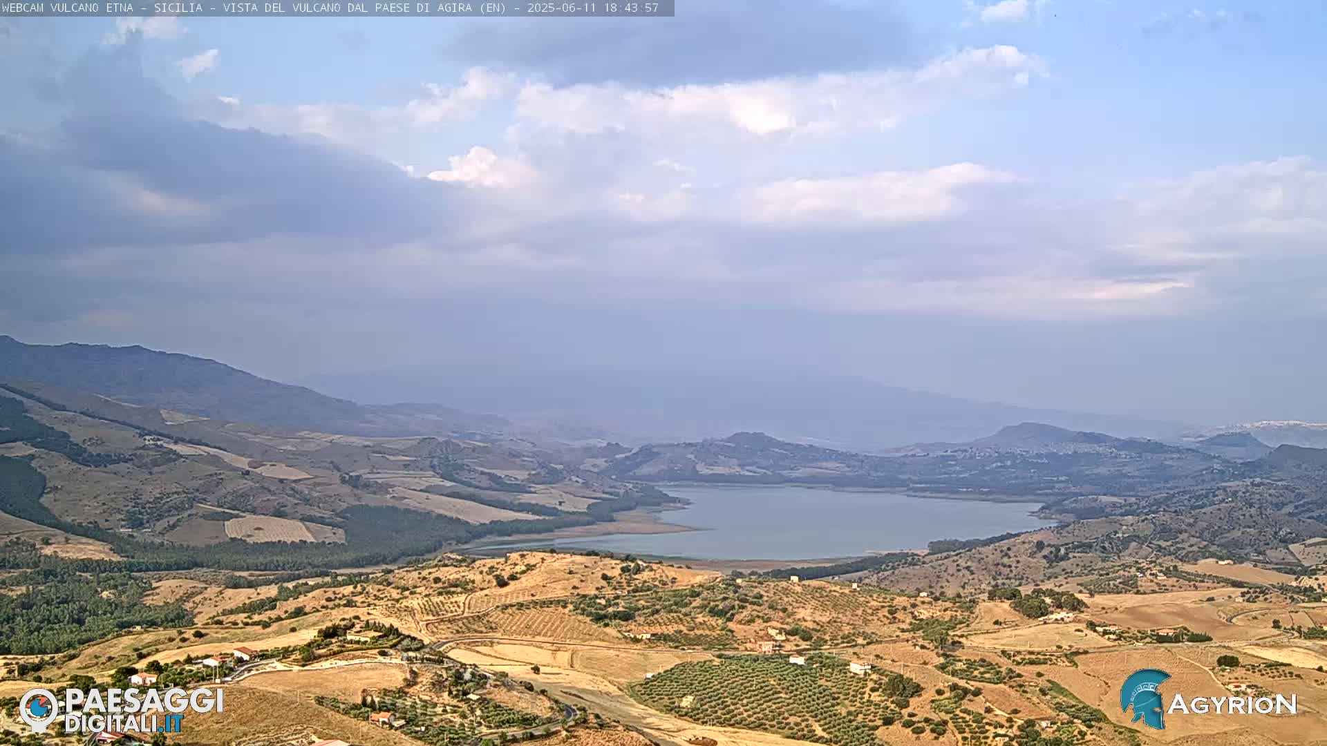 A partly cloudy sky overlooks a vast, dry landscape of rolling hills and farmland surrounding a large lake.