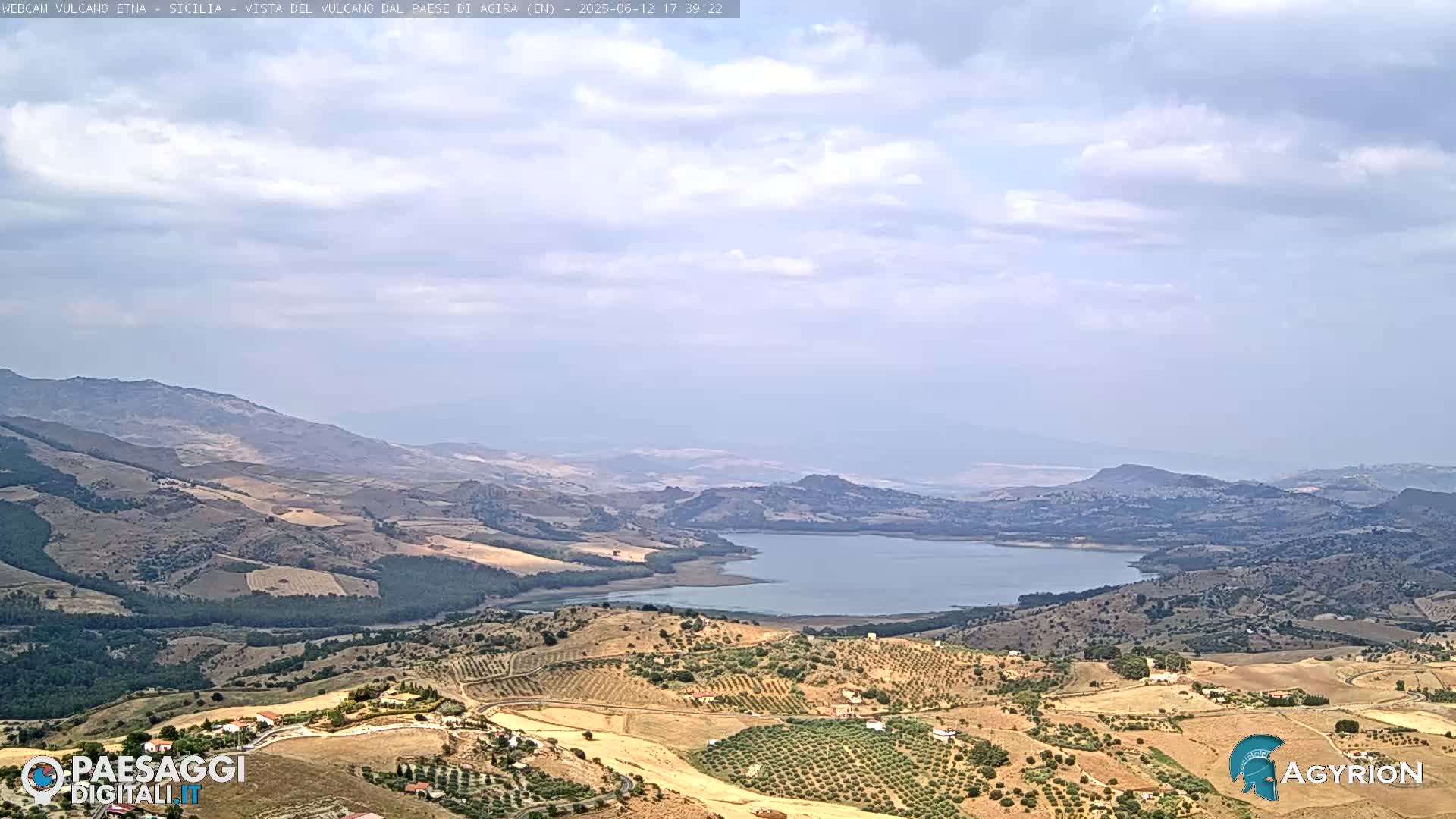 A partly cloudy day reveals a vast landscape of rolling hills, cultivated fields, and a large lake nestled within a valley.