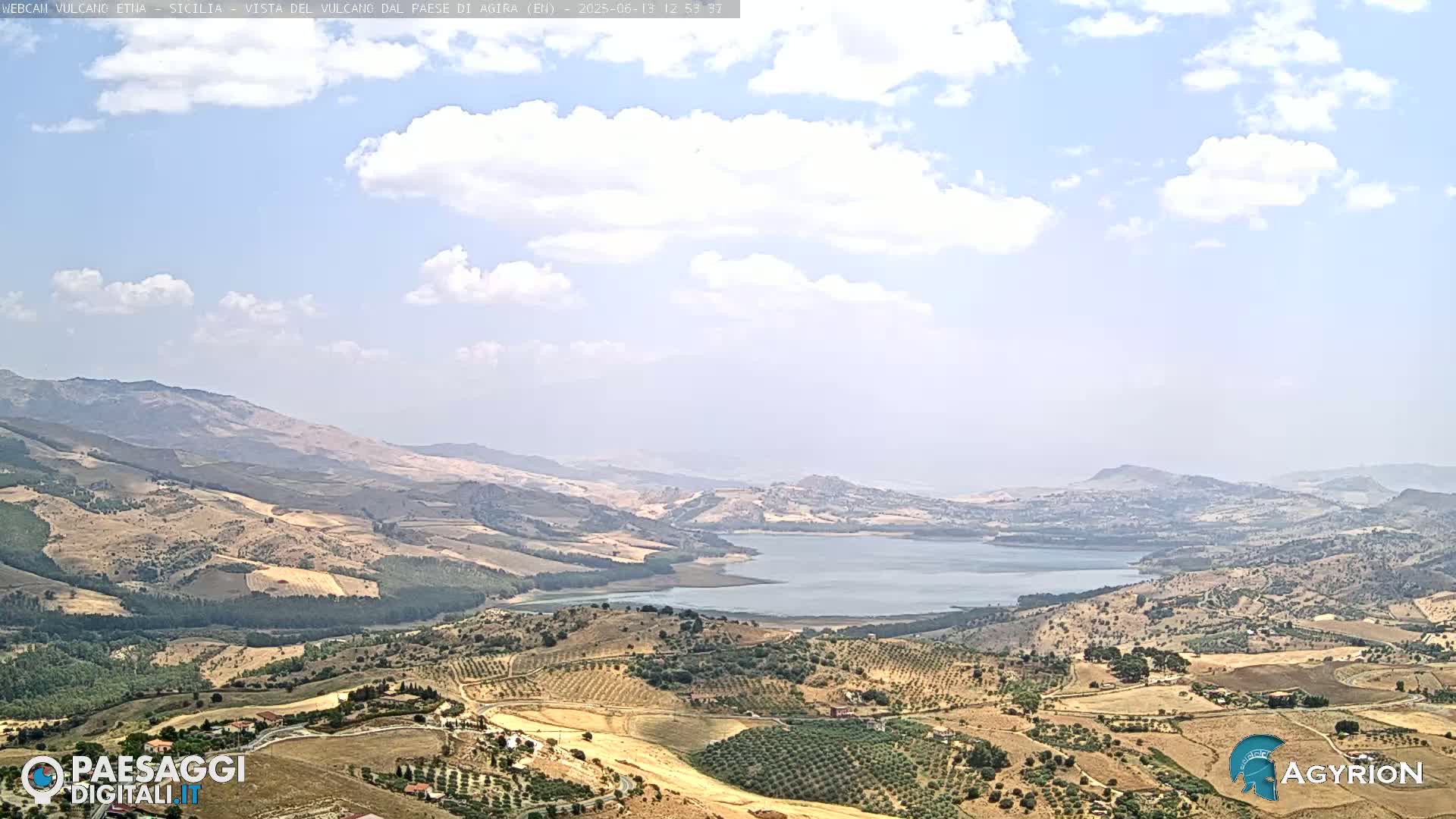 A partly cloudy day reveals a vast landscape of rolling hills and farmland surrounding a calm lake, nestled in a valley between mountains.