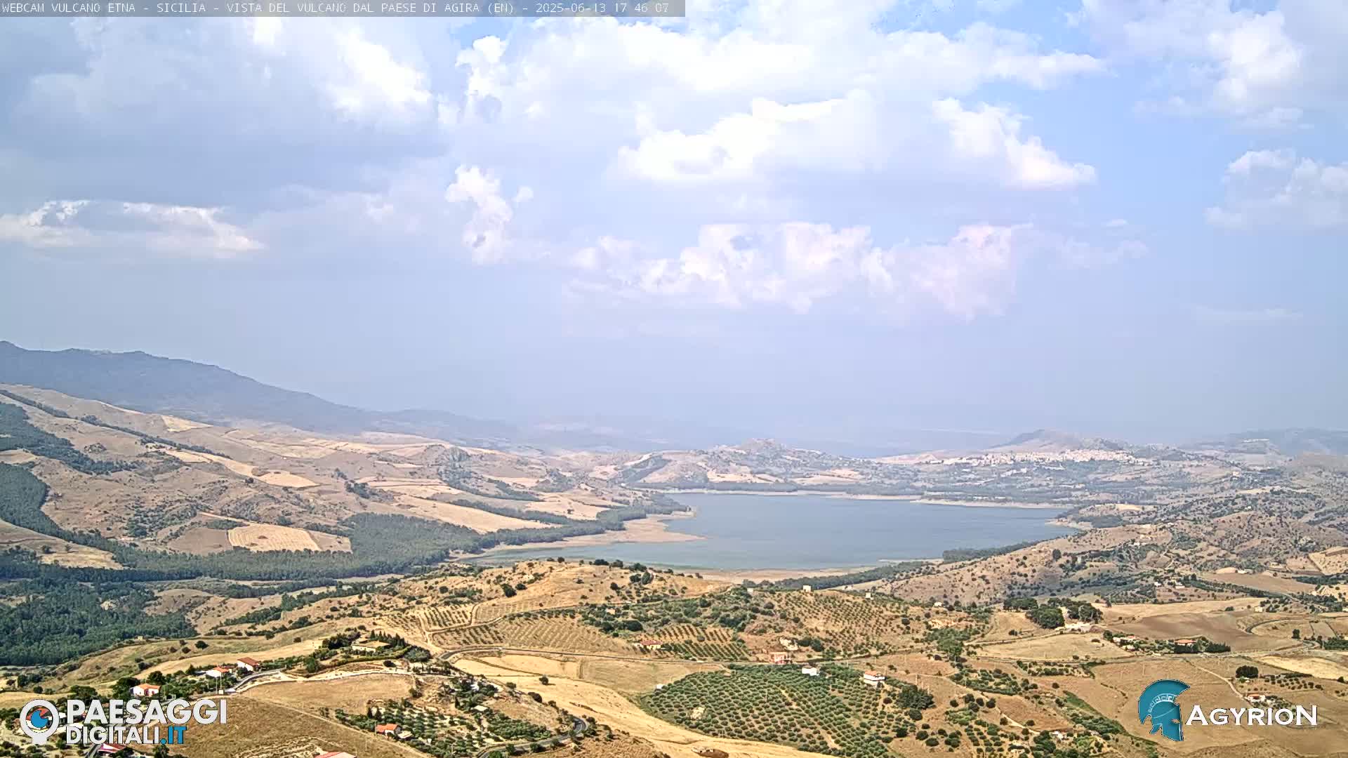 A partly cloudy day reveals a vast, dry landscape with rolling hills, cultivated fields, and a large lake nestled in a valley, all under a mostly clear sky.