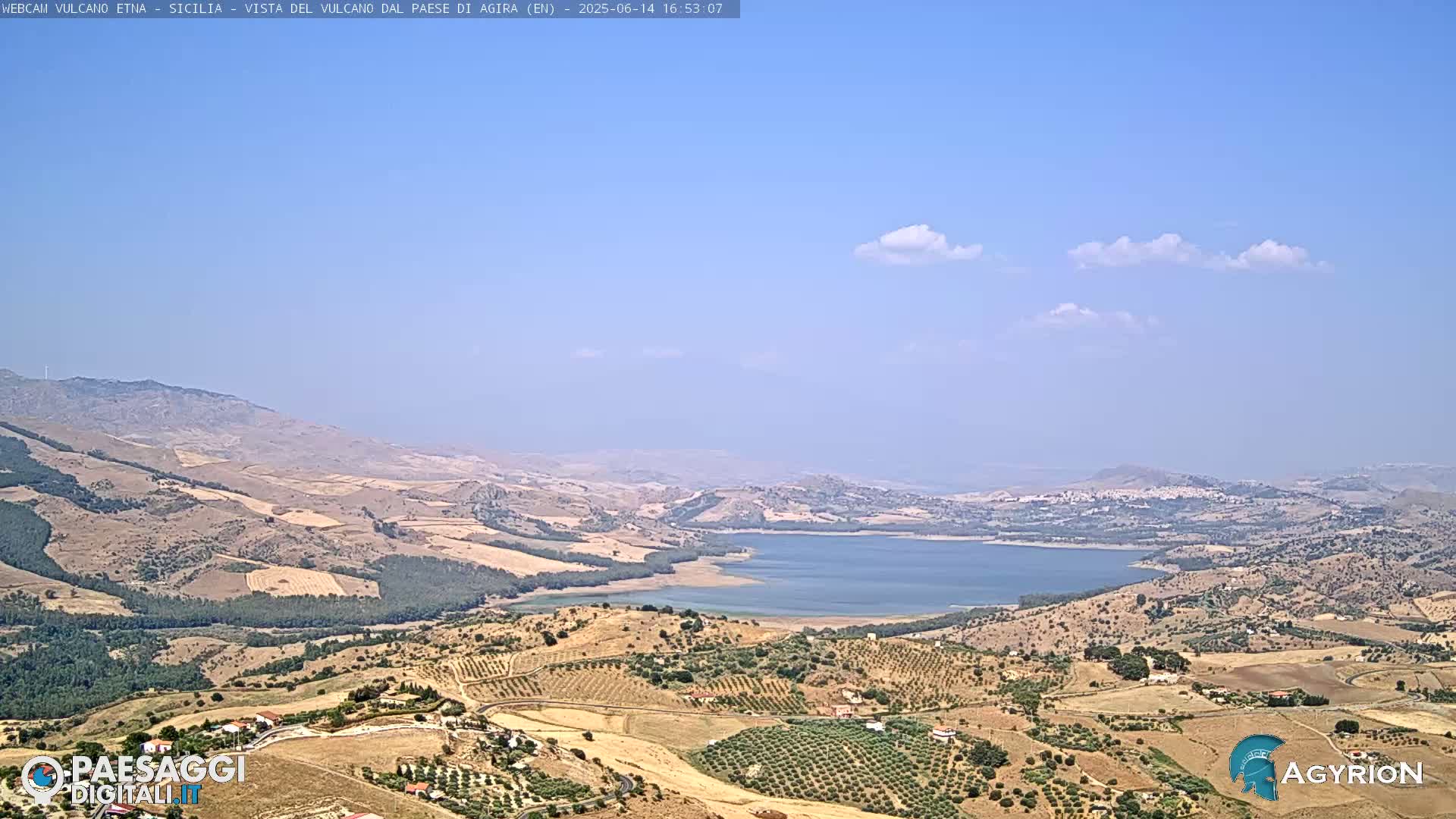 A vast, hilly landscape under a mostly clear blue sky with a few scattered clouds features a large lake nestled in the valley.