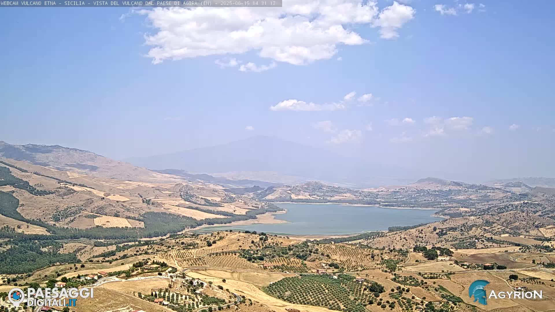A high-angle, long shot of a sun-drenched, hilly landscape with a large lake in the center under a mostly clear sky with a few scattered clouds.