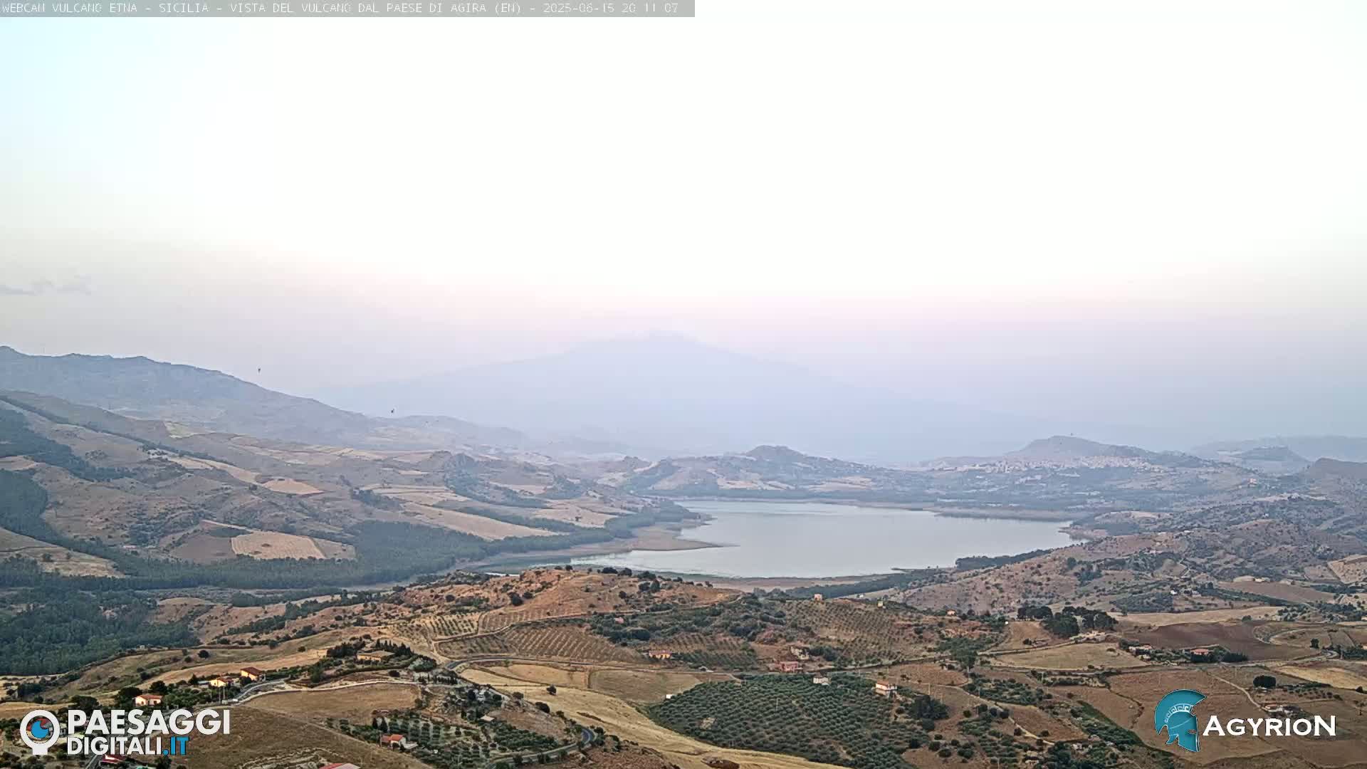 A hazy, high-angle view shows a large lake nestled in a valley between rolling hills and mountains under a light, pale sky.