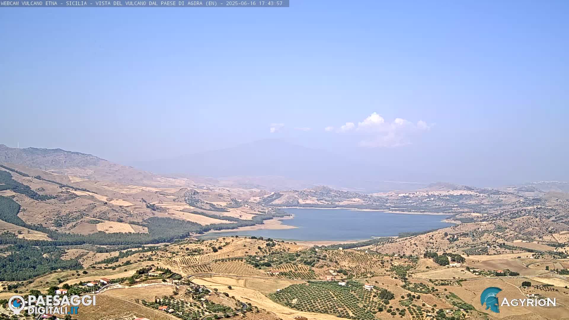 A mostly clear, bright day reveals a hilly landscape with a large lake, cultivated fields, and distant mountains.