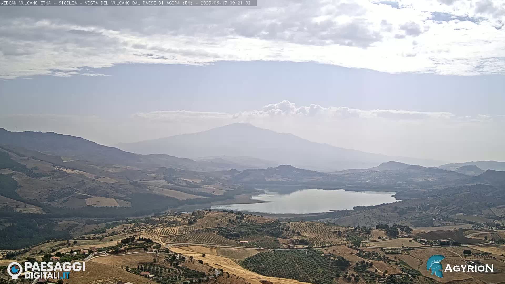 A hazy view shows a large lake nestled in a valley, surrounded by rolling hills and farmland, with a distant, large, hazy mountain in the background under a partly cloudy sky.