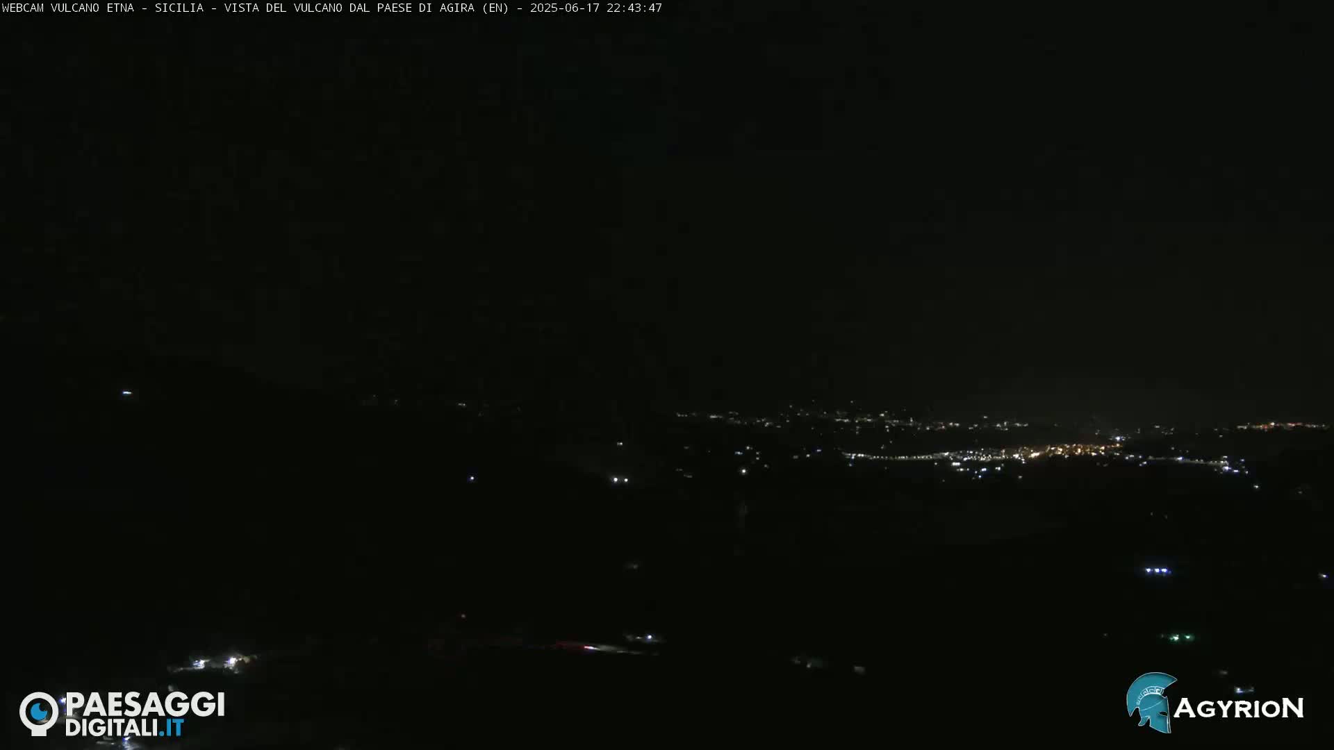 A nighttime long shot of a dimly lit town nestled in a dark landscape under a clear, black sky.
