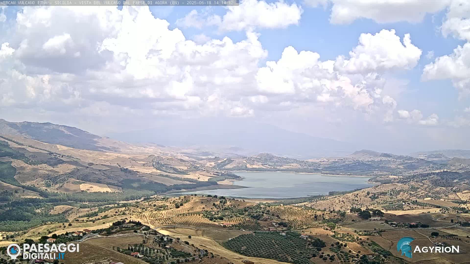 A partly cloudy day reveals a vast, hilly landscape with a large lake in the mid-ground, dotted with olive groves and farmlands.