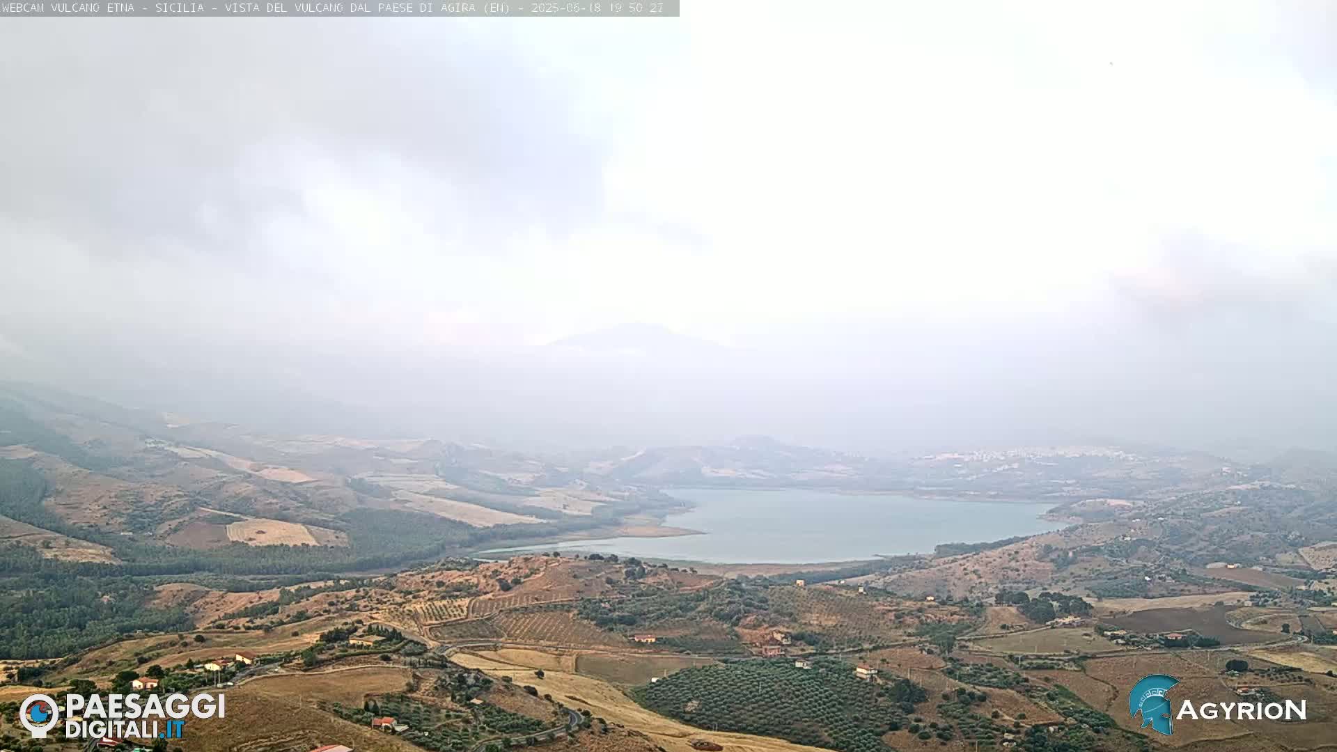 A hazy, high-angle view shows a large lake nestled in a valley of rolling, dry, cultivated hills.