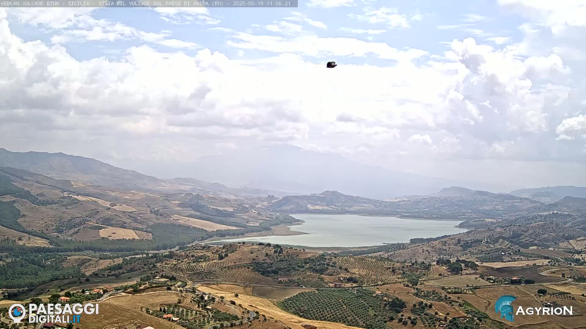 A partly cloudy sky overlooks a vast, hilly landscape with a lake in the middle distance.