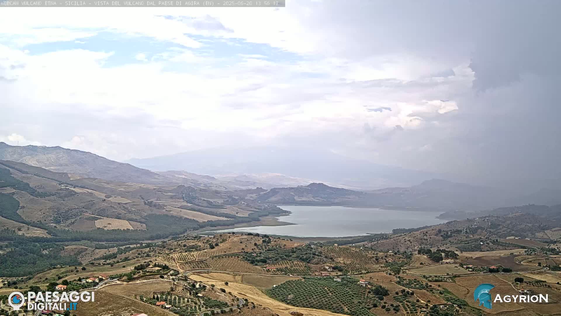 A hilly, dry landscape with cultivated fields and a large lake is visible under a mostly cloudy sky.