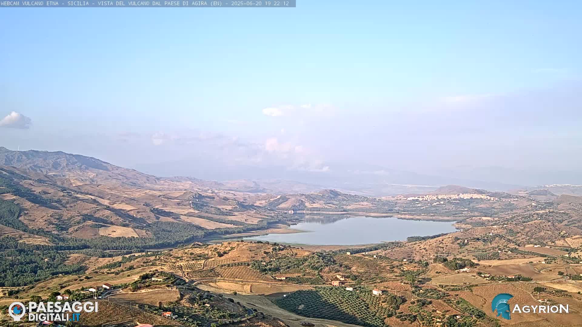 A hazy, sunny day reveals a vast, hilly landscape with a calm lake nestled among dry, cultivated fields and distant mountains.