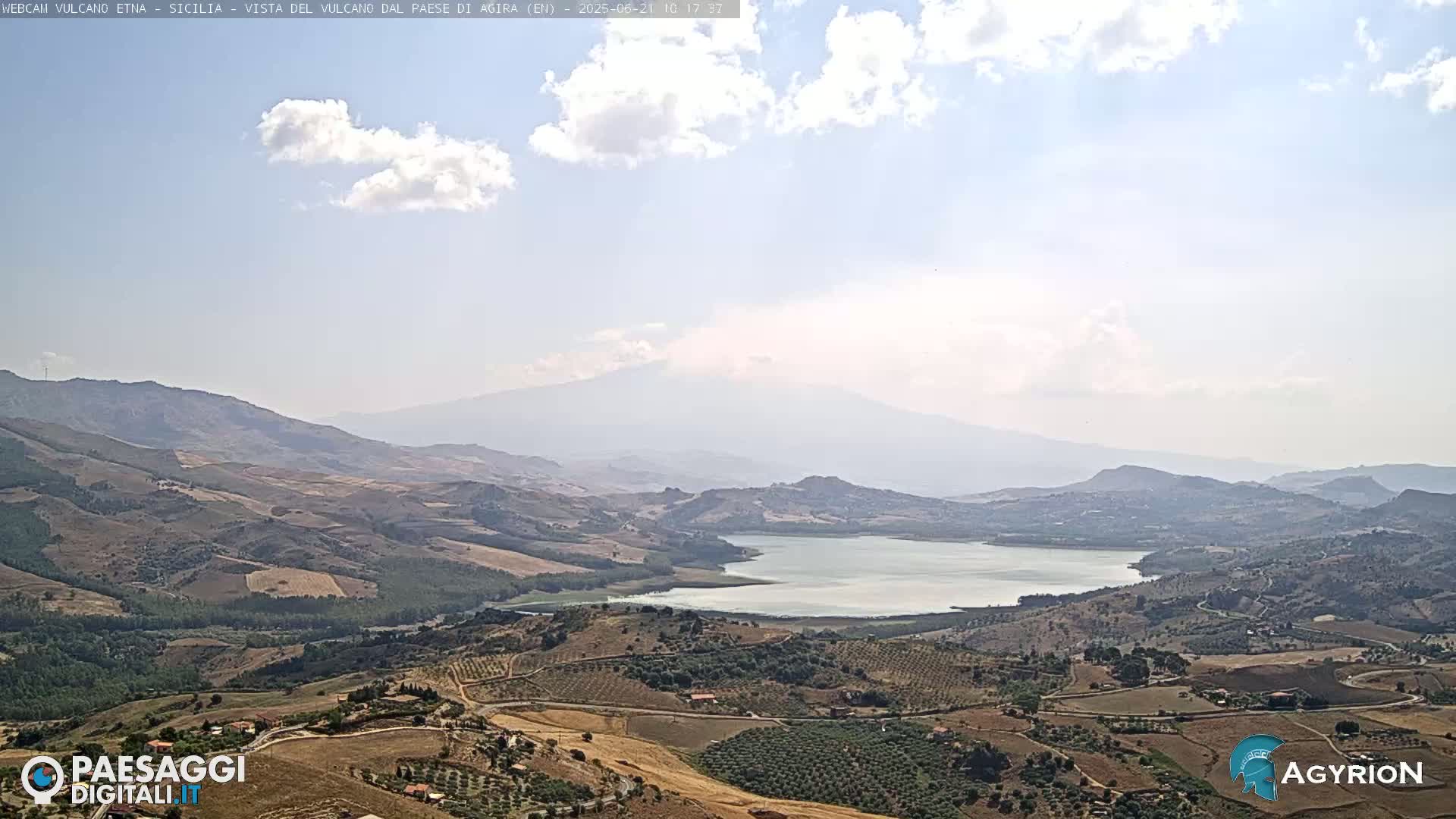 A partly cloudy sky overlooks a vast, hilly landscape with a lake in the middle distance and a large volcano visible in the far background.
