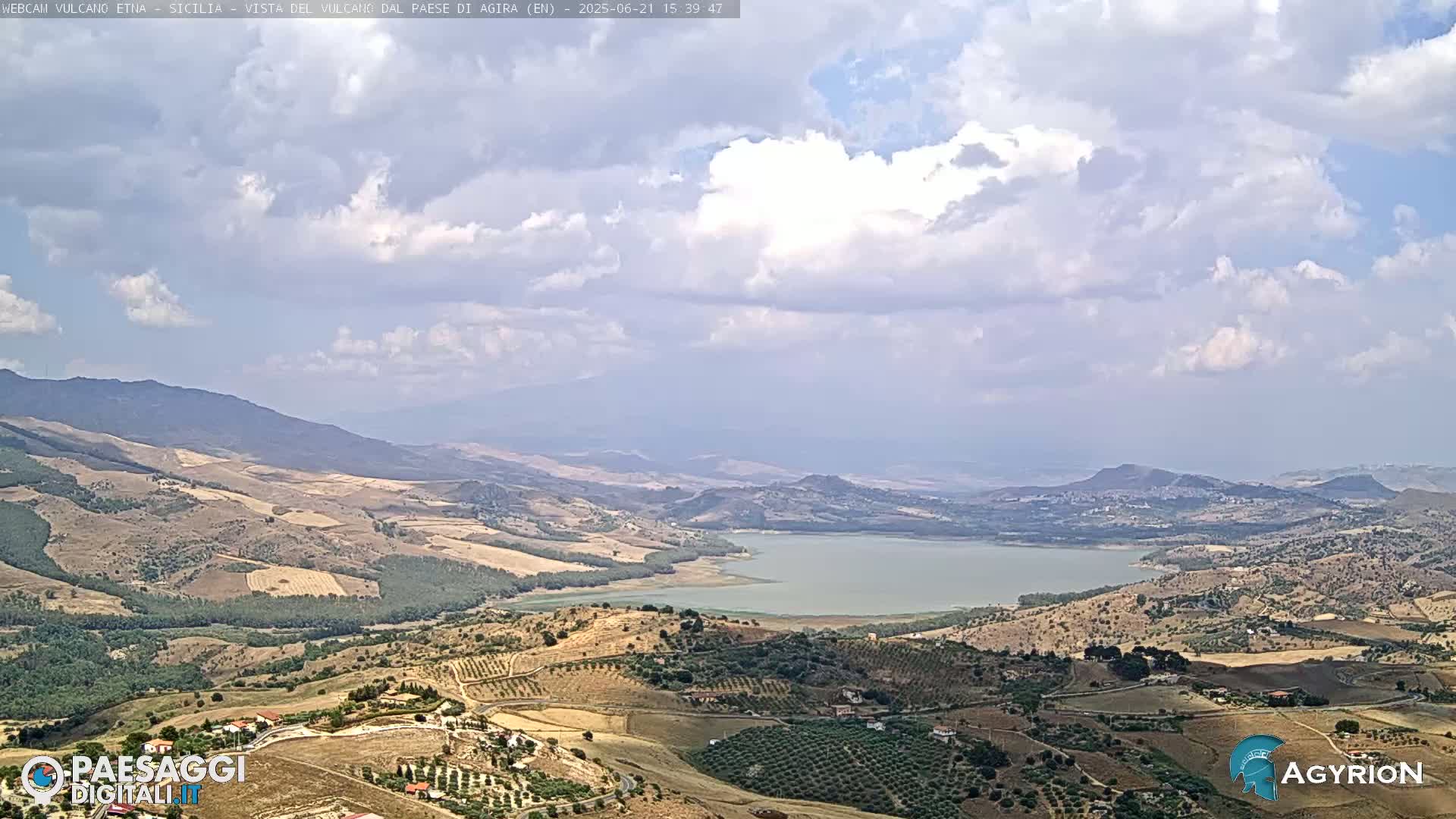 A partly cloudy sky overlooks a vast, hilly landscape with a large lake nestled in the valley below.
