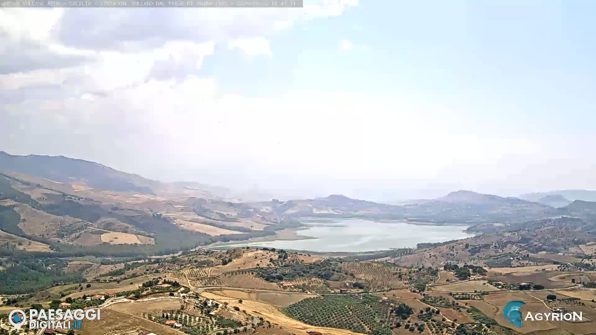 A hazy, partly cloudy day reveals a sprawling landscape of rolling hills and farmland surrounding a large lake, nestled among mountains in the distance.