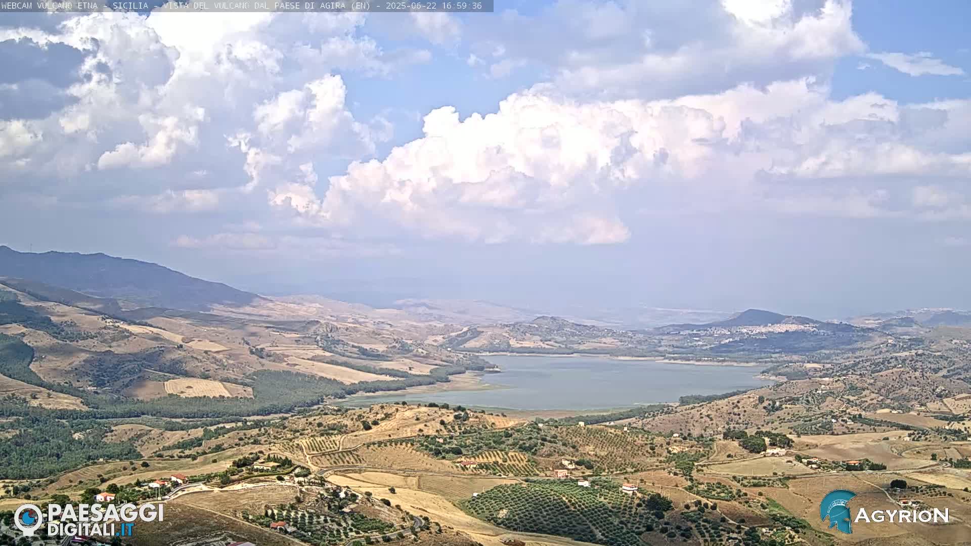 A partly cloudy day reveals a vast, hilly landscape dotted with olive groves and a large lake, with mountains in the distance.