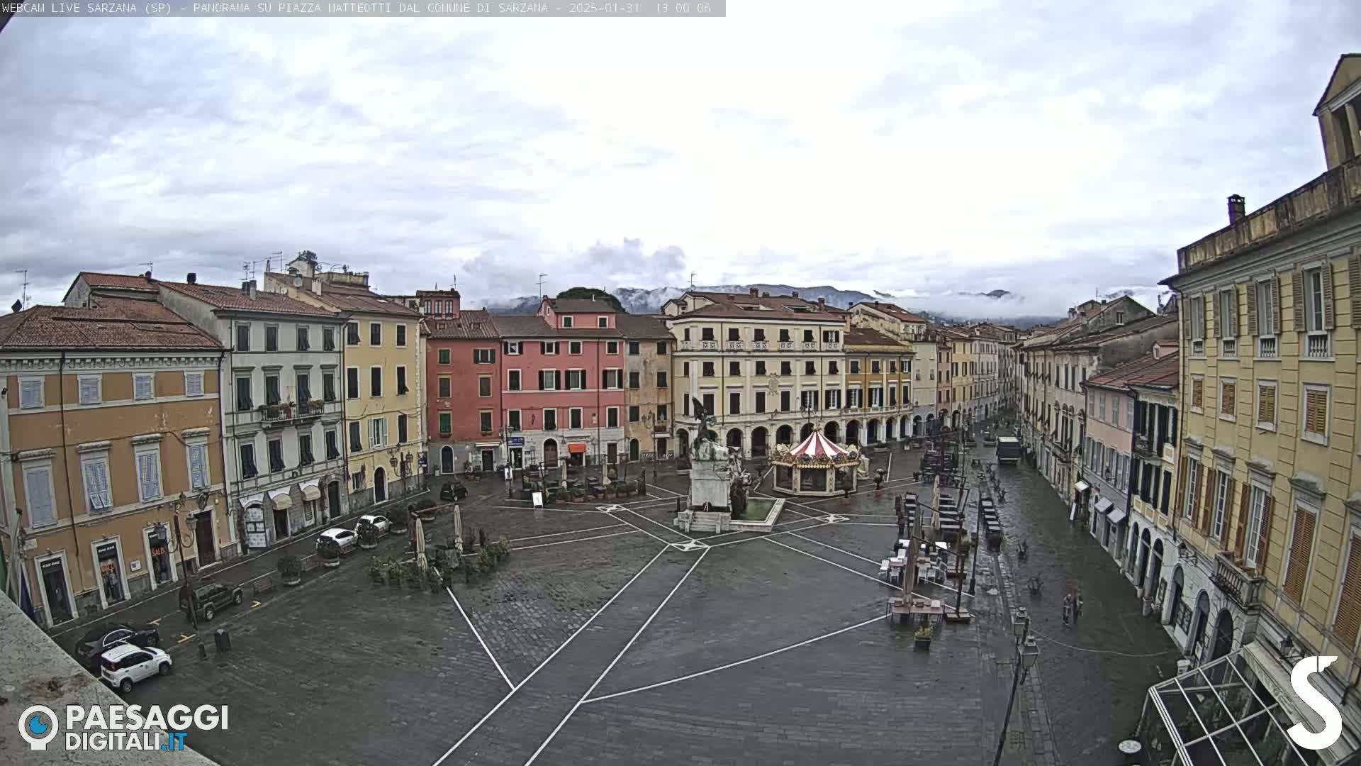 A wet town square with several buildings surrounding it, a monument in the center, and distant mountains under a cloudy sky.