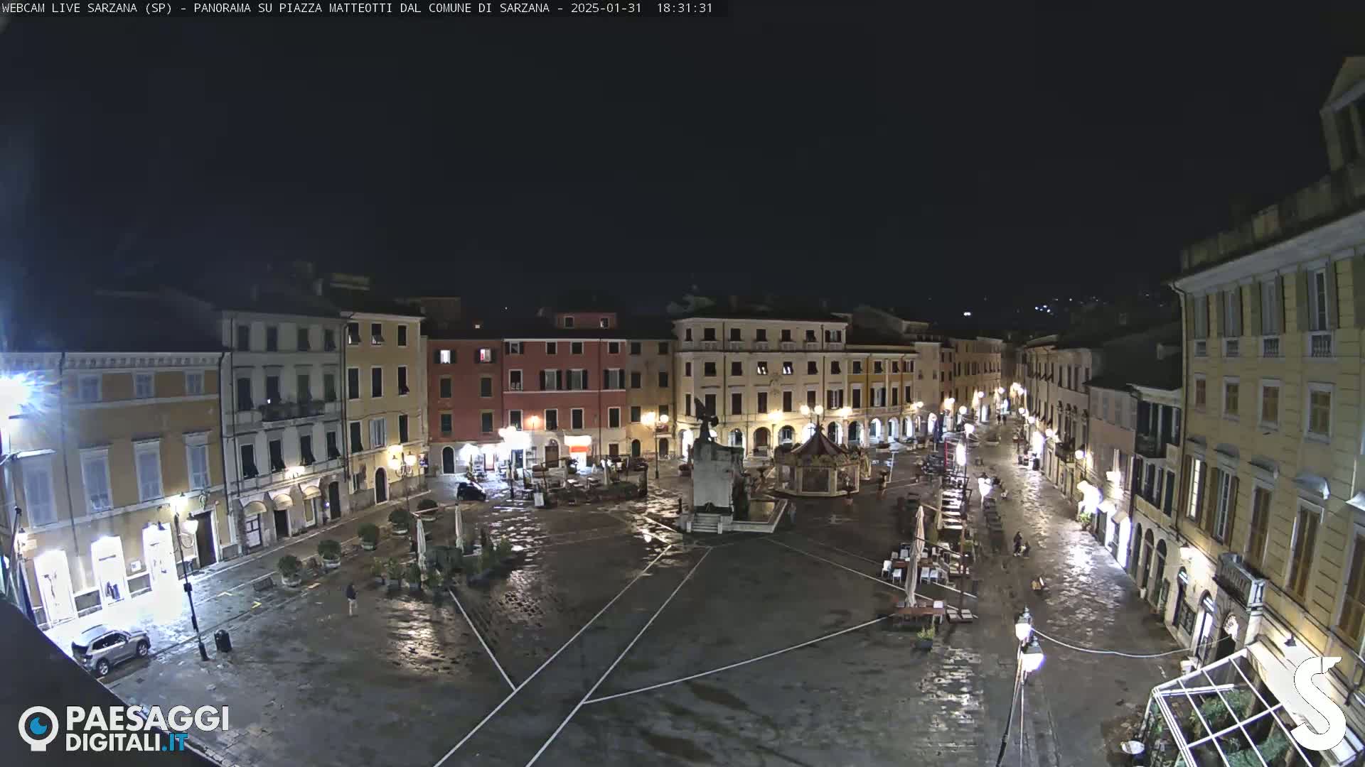 A wet, nighttime view of a town square with several buildings surrounding it, lit by streetlights.