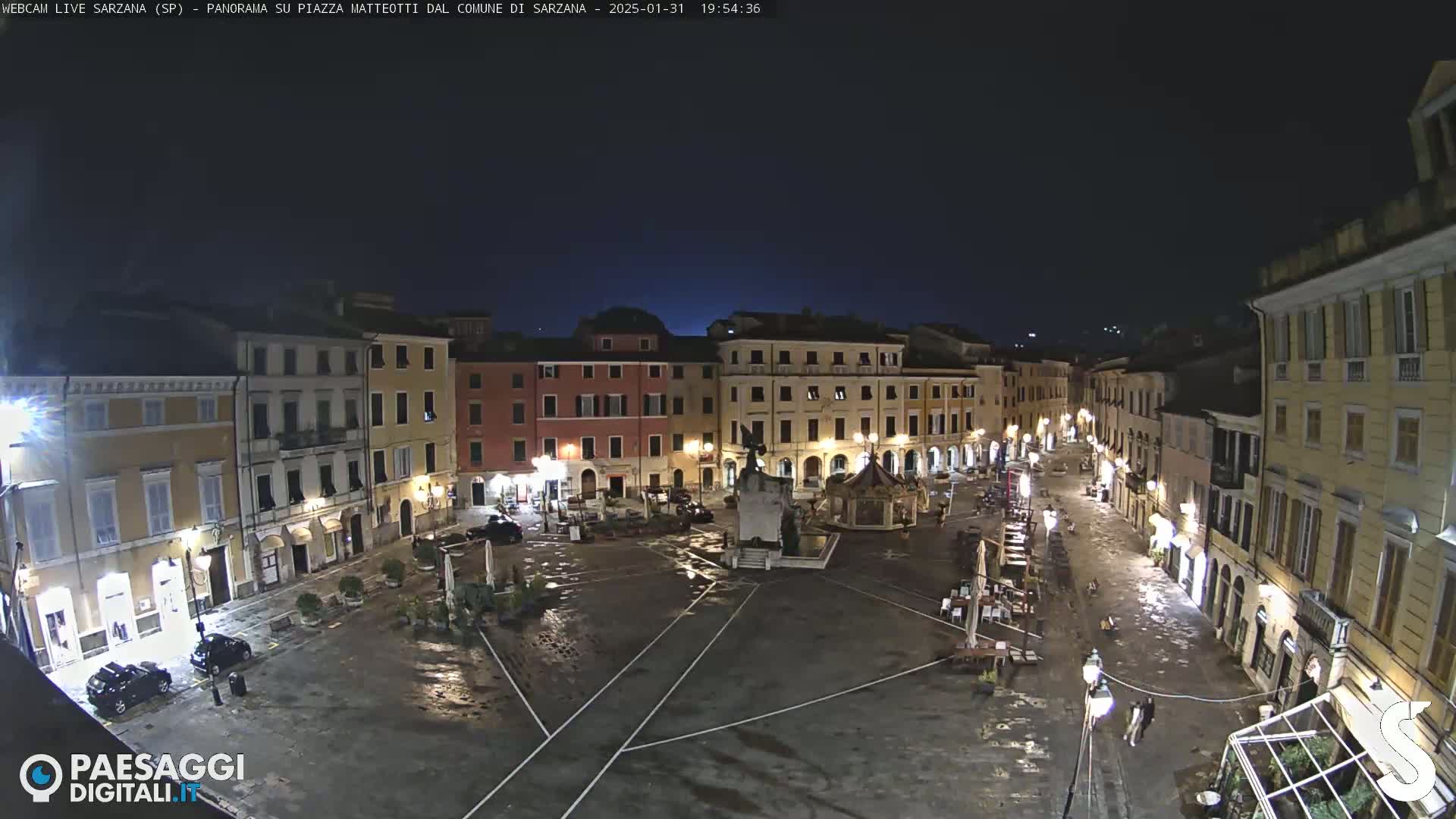 A nighttime view of a mostly empty town square surrounded by illuminated buildings, under a dark sky.