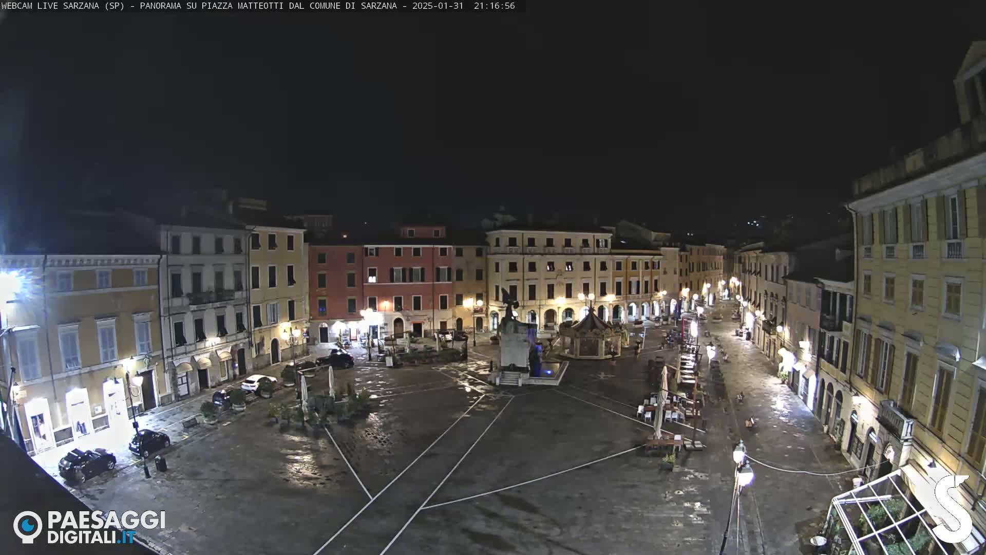 A wet, nighttime view of a town square surrounded by buildings, with a monument and sparse outdoor seating visible.