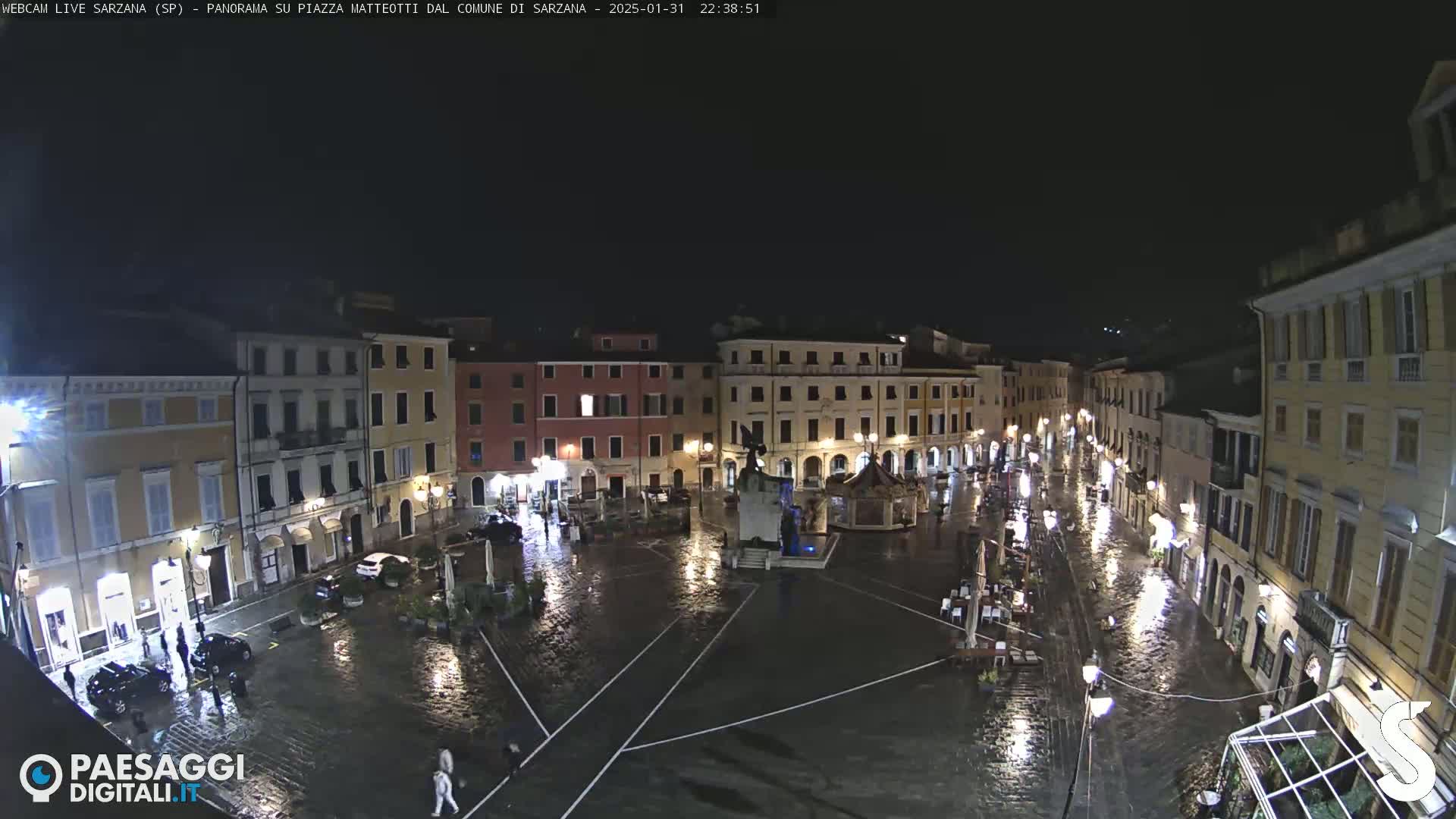 A rain-soaked town square at night, surrounded by illuminated buildings, is seen from an elevated perspective.