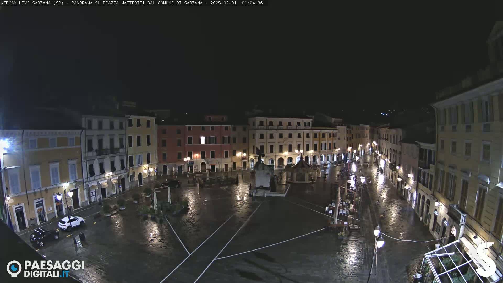 A rain-soaked town square at night, surrounded by illuminated buildings.