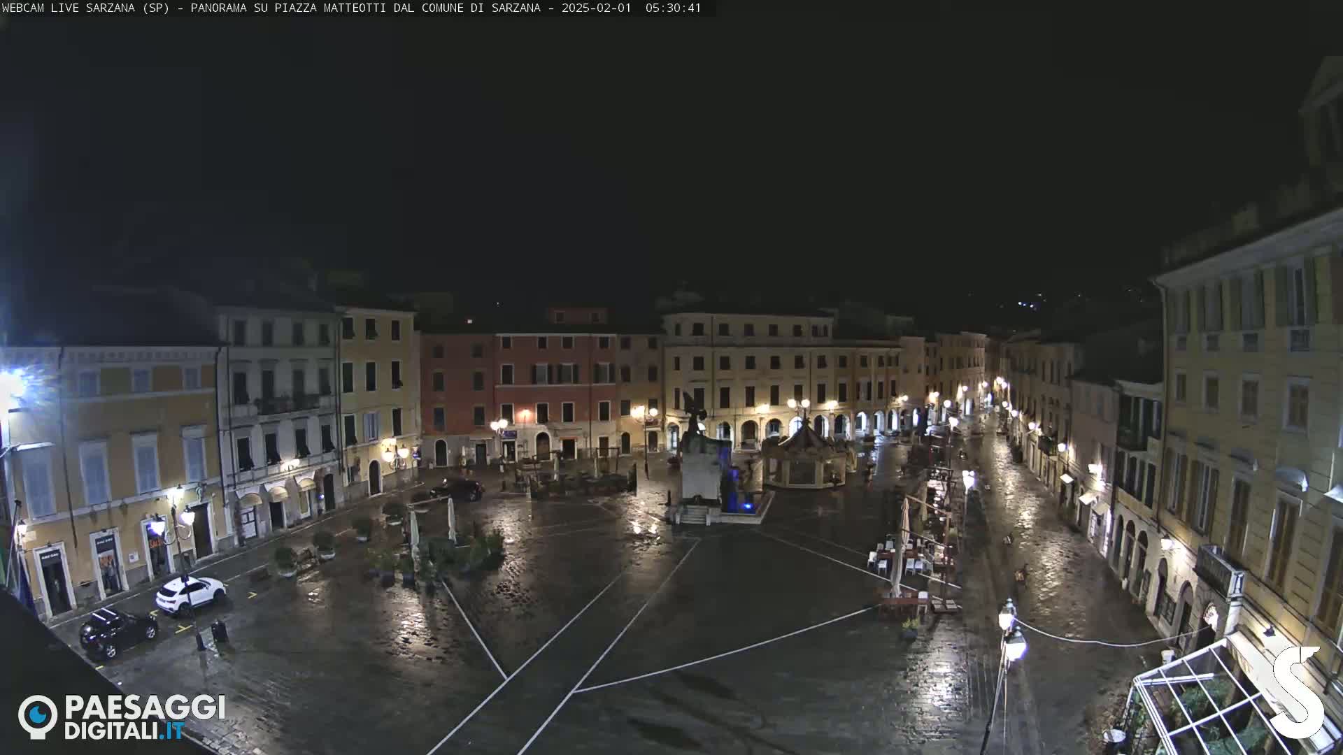 A rain-slicked town square at night, surrounded by illuminated buildings, features a central monument and a few parked cars.