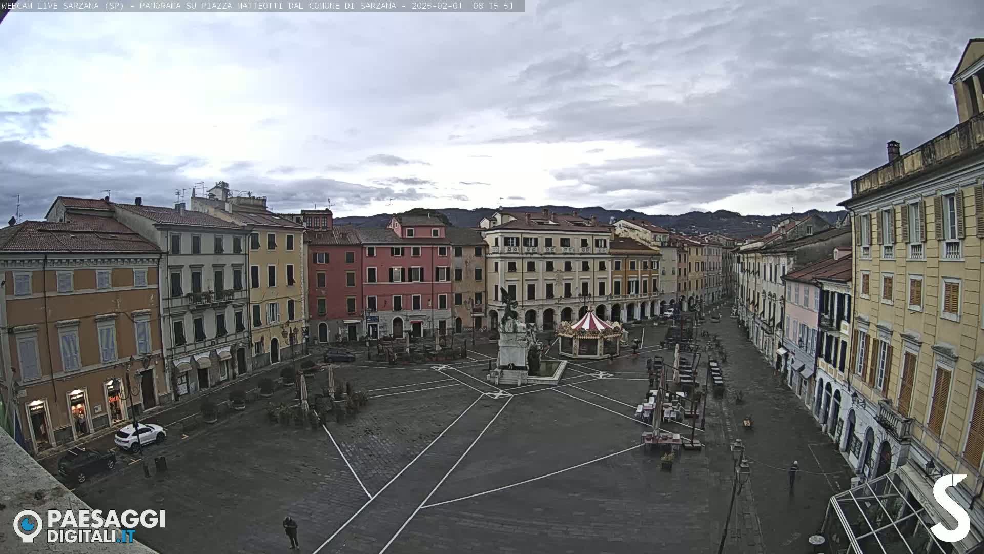 A mostly empty town square with multi-story buildings surrounding it under an overcast sky.