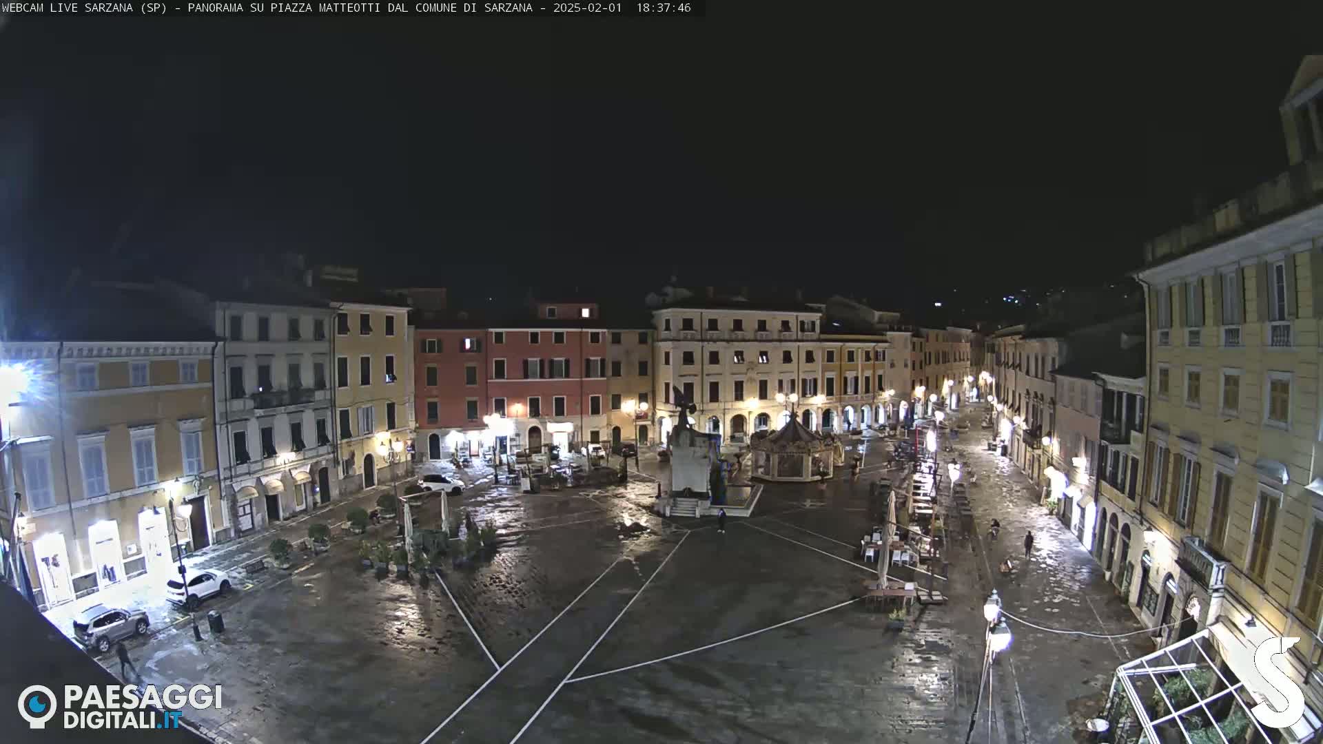 A wet, nighttime view of a town square surrounded by buildings, with a few cars and people sparsely visible.