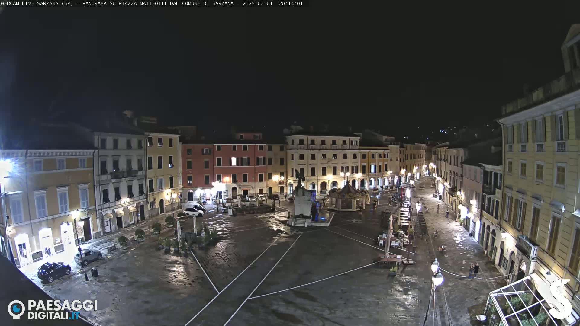 A wet, nighttime view of a town square surrounded by illuminated buildings.