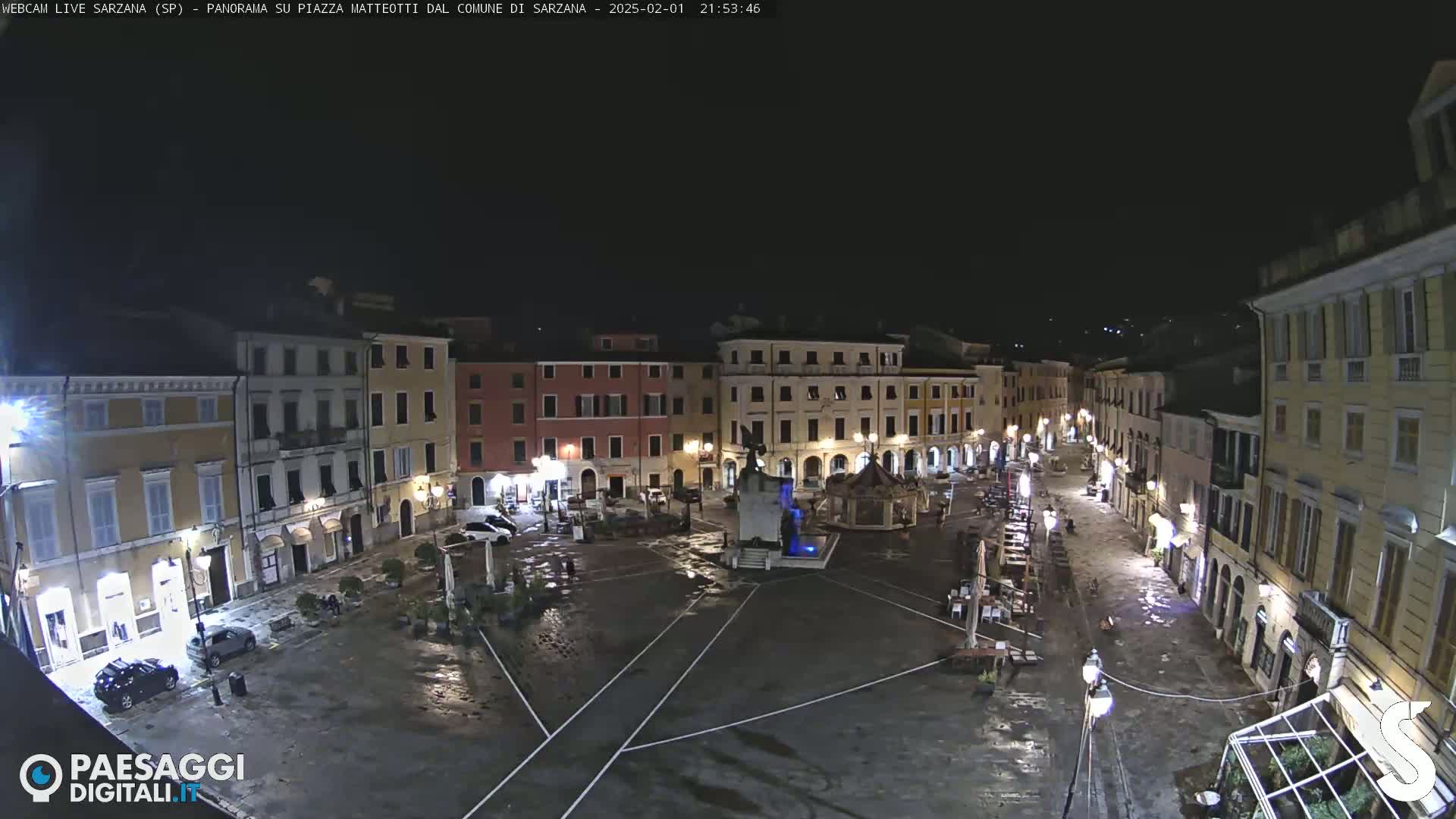 A wet, nighttime view of a town square with buildings surrounding it, a central fountain, and a few cars parked nearby.