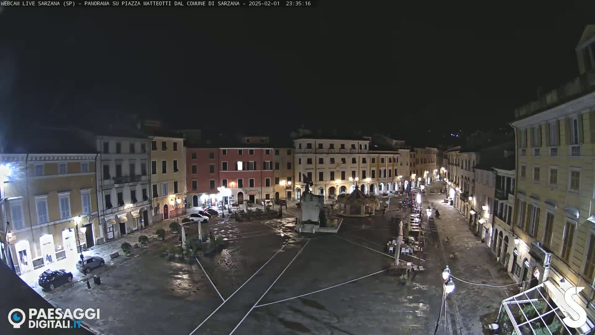 A wet, empty town square at night, surrounded by illuminated buildings.