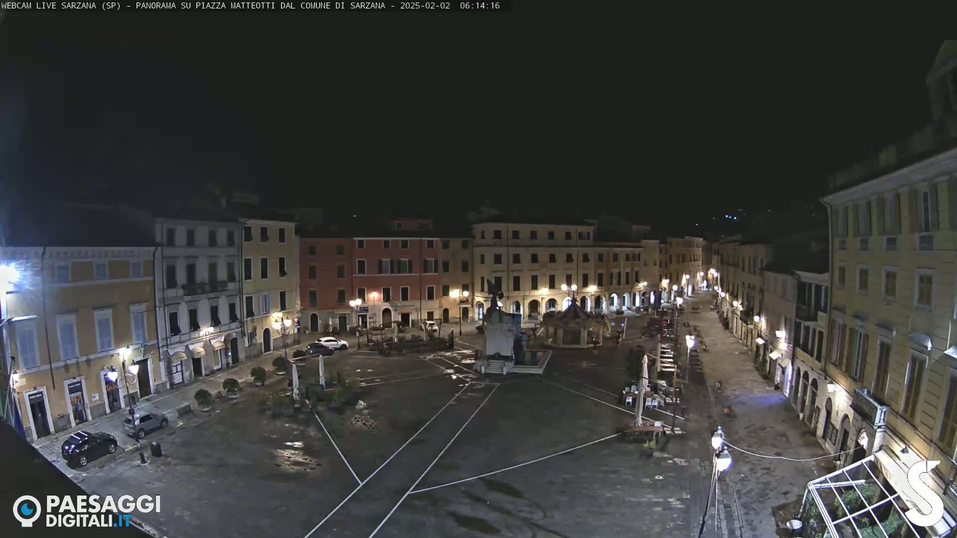 A nighttime, mostly empty town square, surrounded by buildings with illuminated windows, and a central fountain.