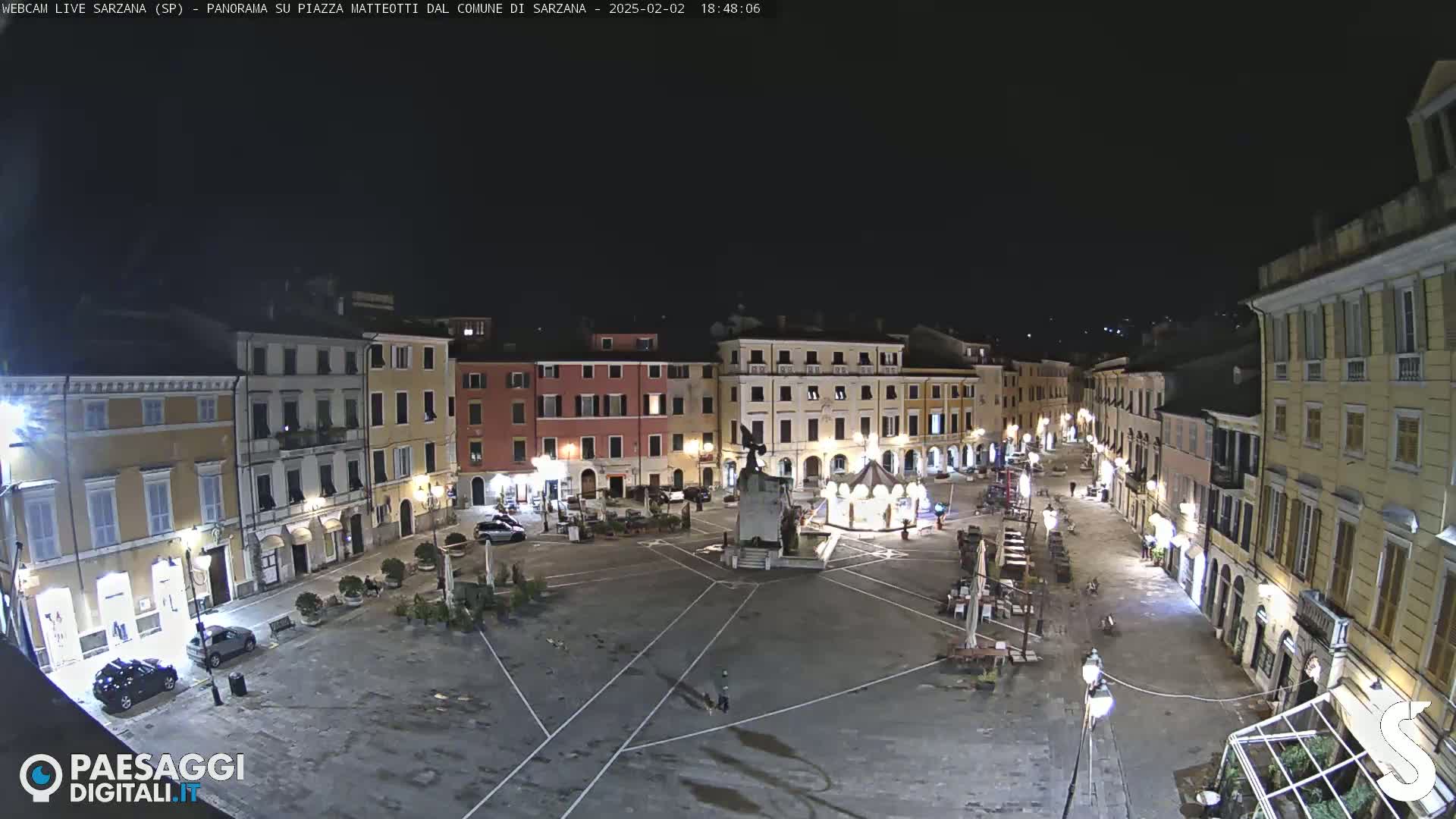 A nighttime, overhead view of a town square with several buildings illuminated, a central monument, and a few people visible.