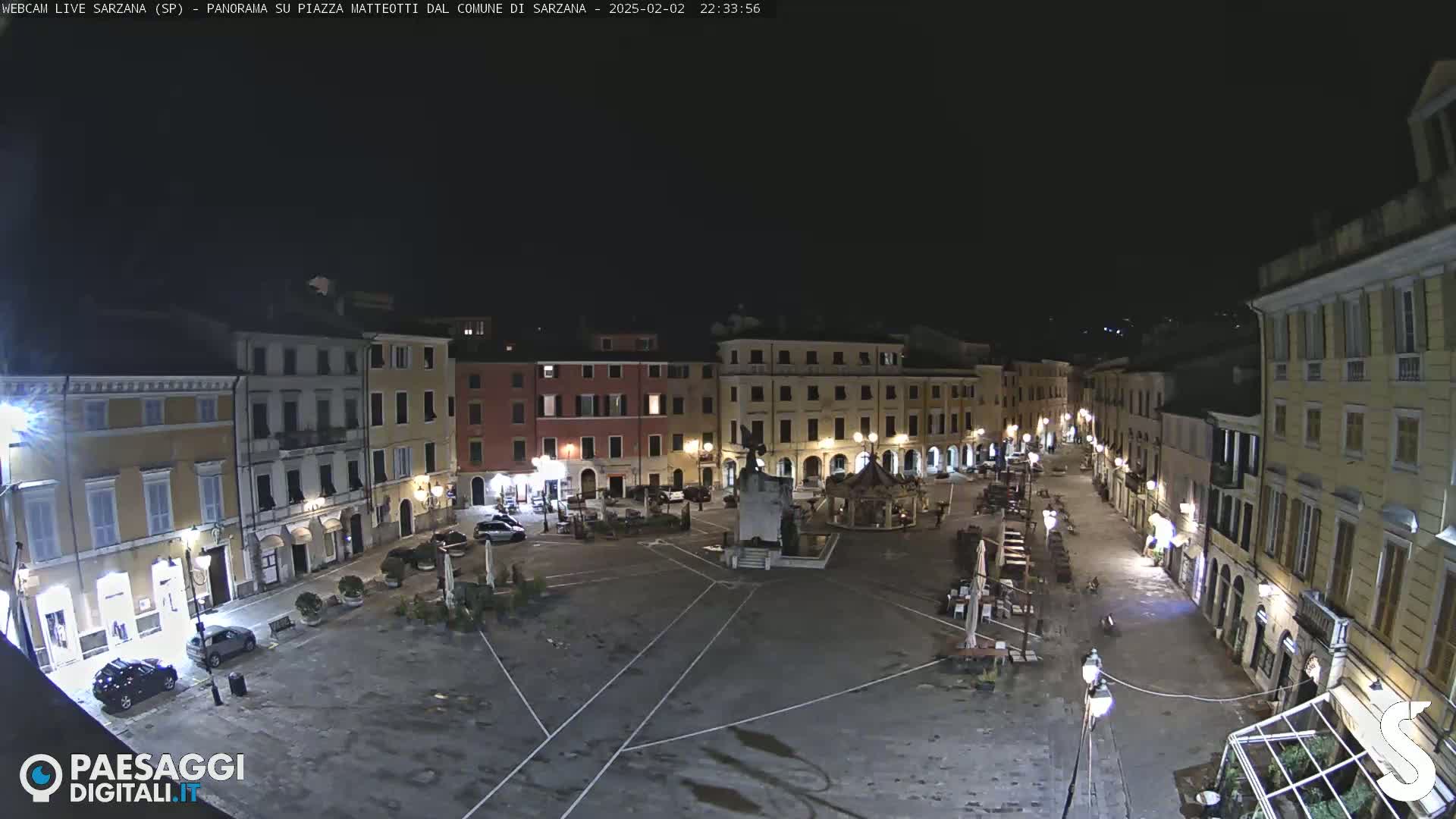 A nighttime, aerial view of a town square with several buildings surrounding it, and a few cars parked.