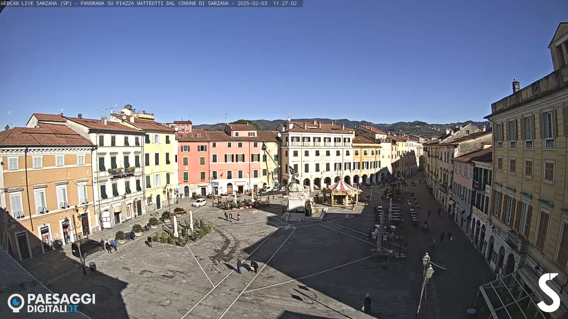 A sunny, clear day overlooks a large town square surrounded by multi-story buildings, with a few people and some outdoor seating visible.