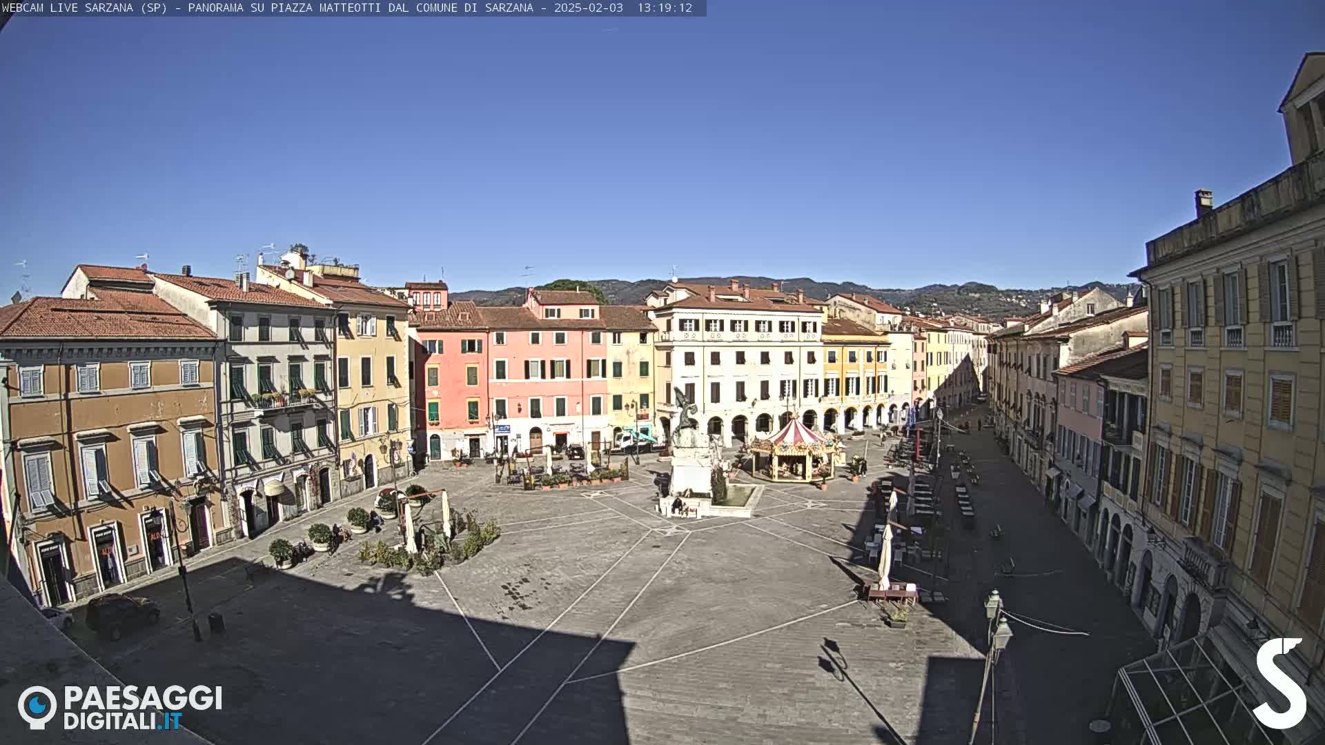 A large, mostly empty town square with multi-story buildings surrounding it under a clear, sunny sky.