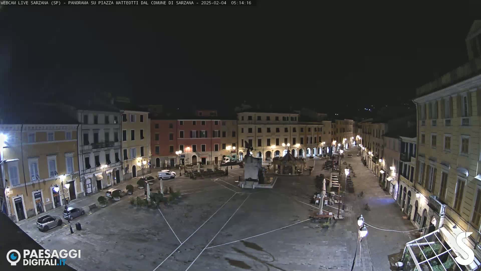 A nighttime, high-angle view of a mostly empty town square, surrounded by illuminated buildings.