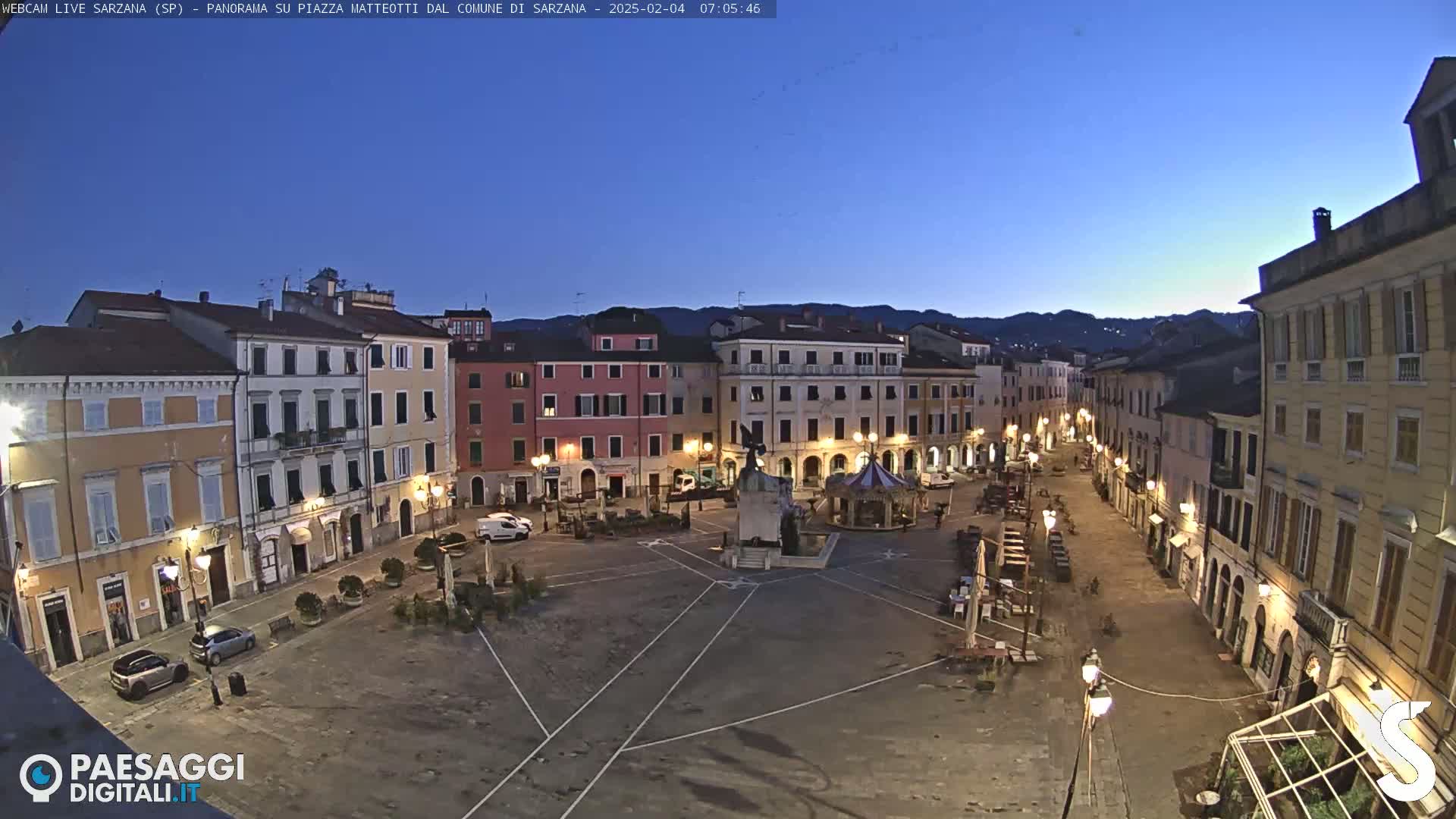 A mostly empty town square, surrounded by multi-story buildings, is shown at twilight under a clear, dark-blue sky.