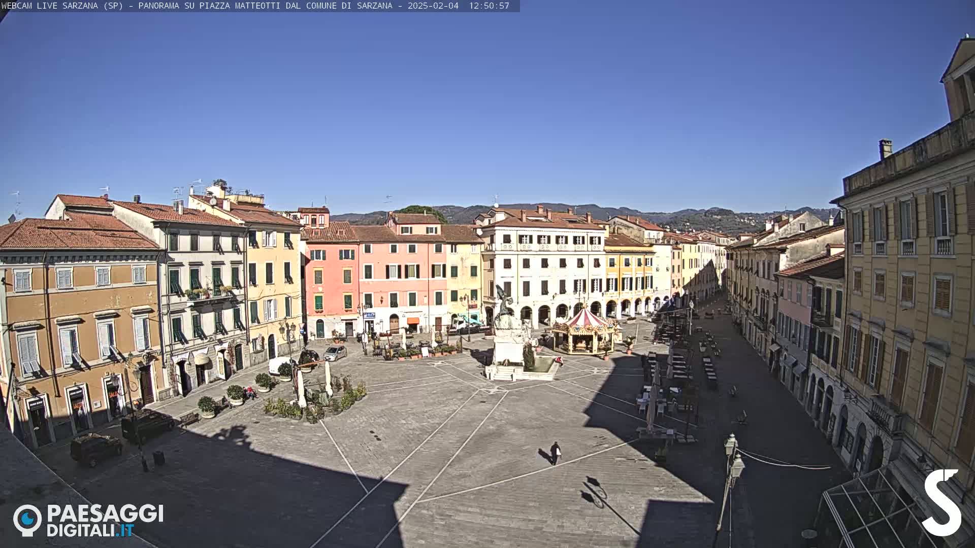 A mostly empty town square with pastel-colored buildings surrounding it on a sunny day.